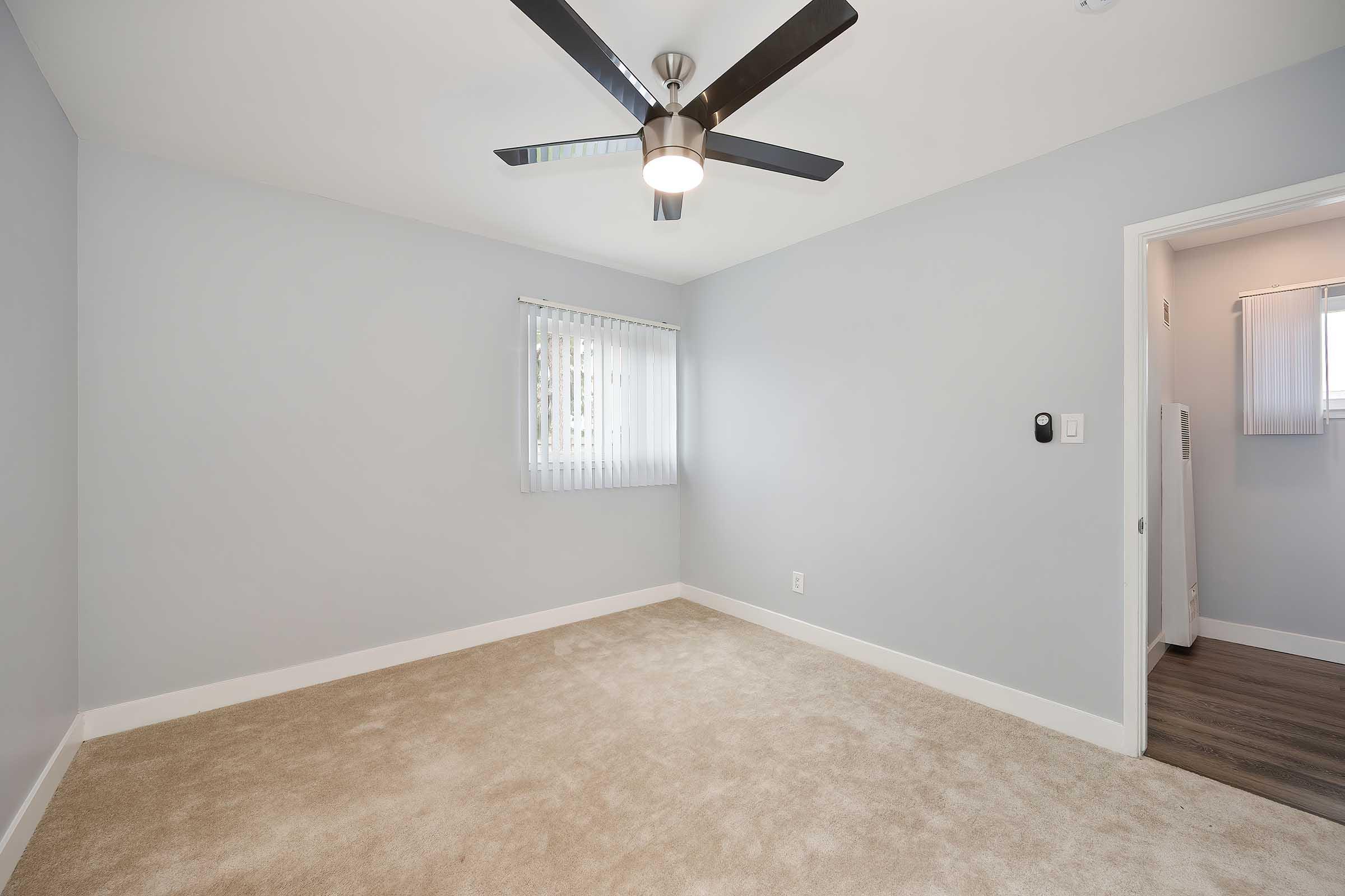 Empty room with light gray walls and beige carpet. A ceiling fan with five blades is installed, and there's a window with vertical blinds allowing natural light. A door leads to another room, and a wall-mounted thermostat is visible. The overall feel is modern and minimalistic.