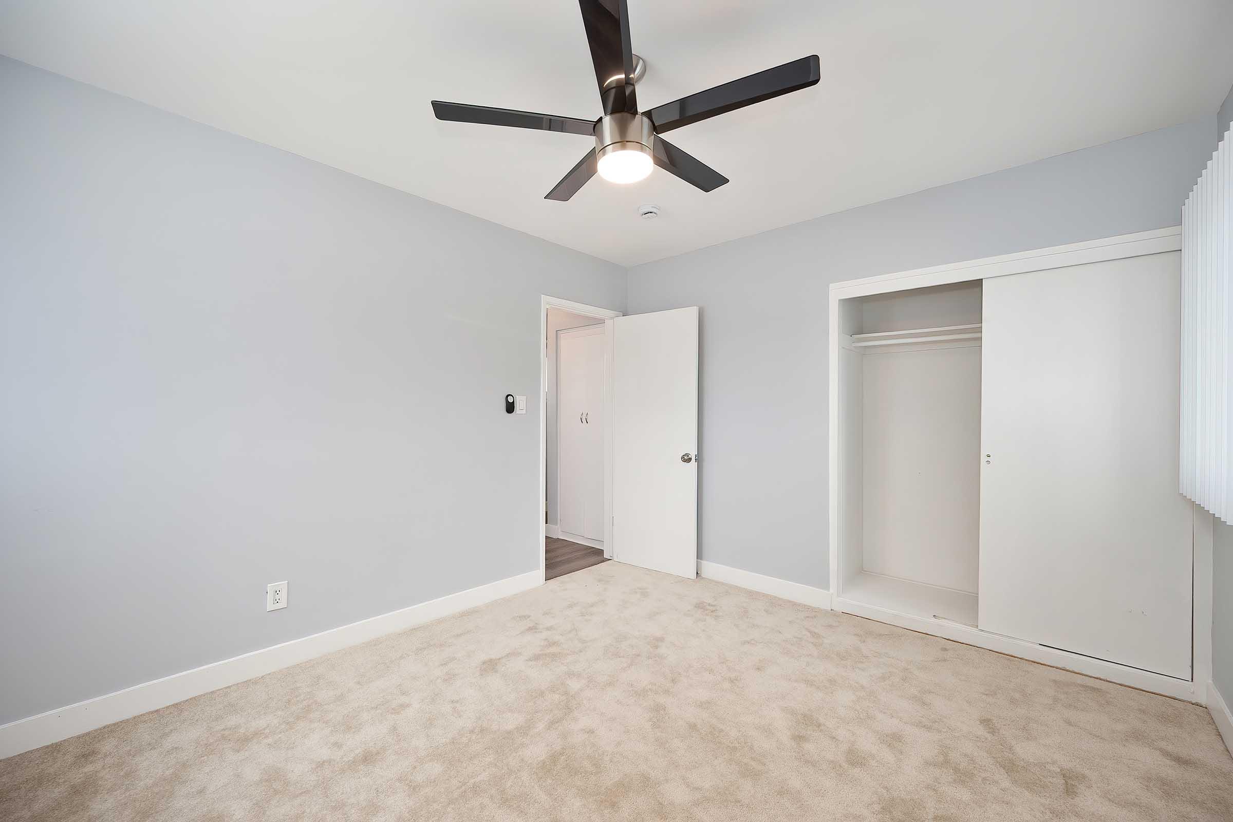 A spacious, minimalist bedroom with light gray walls and a ceiling fan. The room features plush beige carpet, a closet with sliding doors, and a door leading to another area. Natural light streams in through a window with vertical blinds, creating a calm and inviting atmosphere.