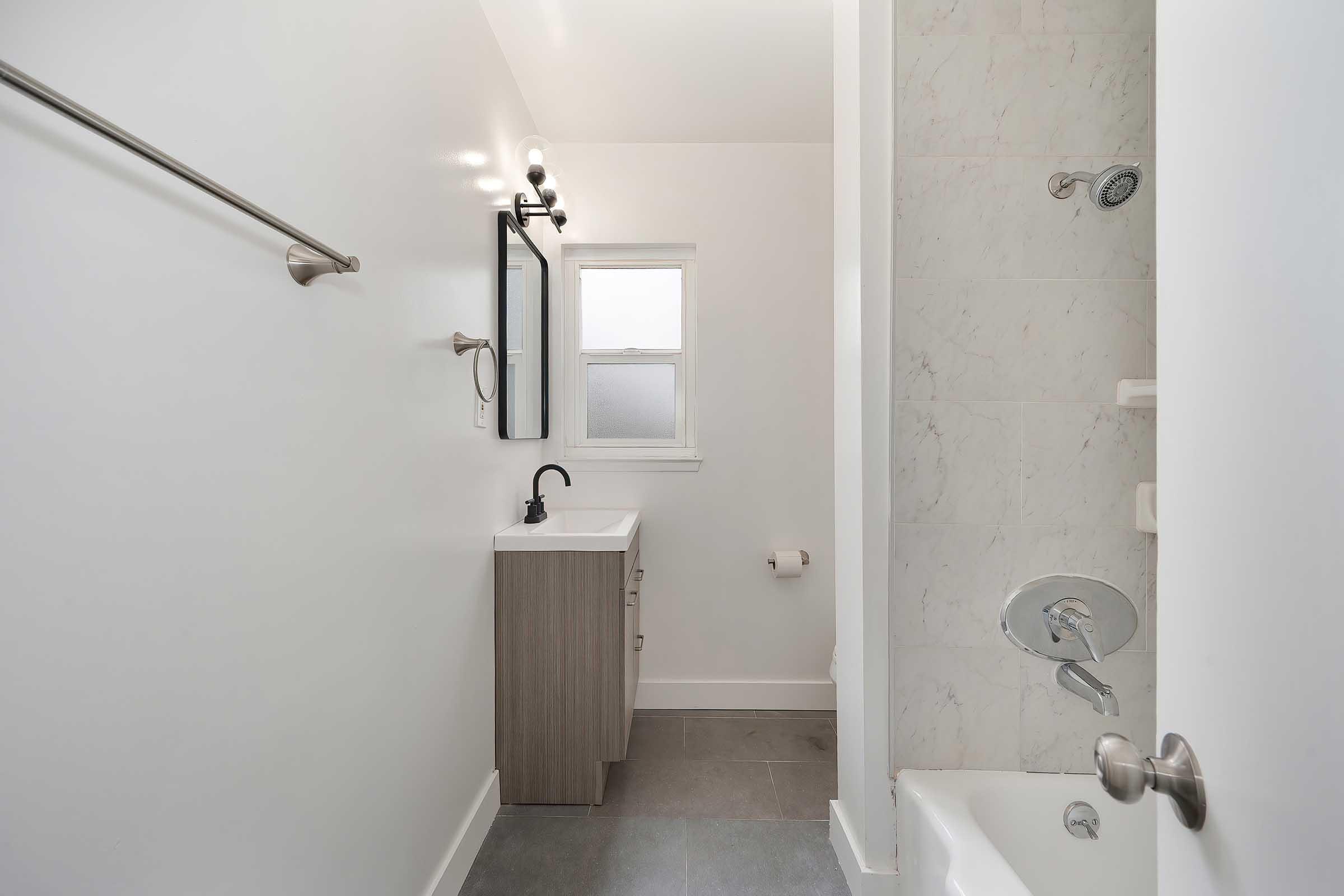 A clean, modern bathroom featuring a white wall, a sleek sink with a black faucet, a wall-mounted mirror, and a shower tub combination. Natural light enters through a window, and there's a towel bar on the wall. The flooring is gray tiles, creating a fresh and minimalist atmosphere.