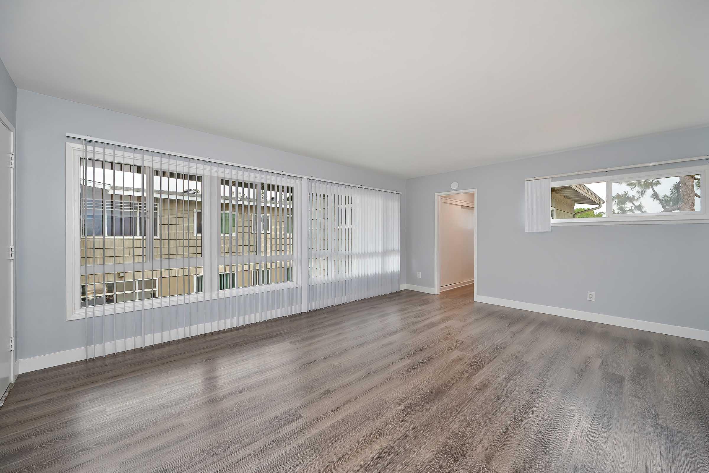 A spacious, empty room featuring light gray walls and brown wood-like flooring. Large windows with vertical blinds allow natural light to fill the space. There is a doorway leading to another room on the right, and the overall ambiance is clean and modern.