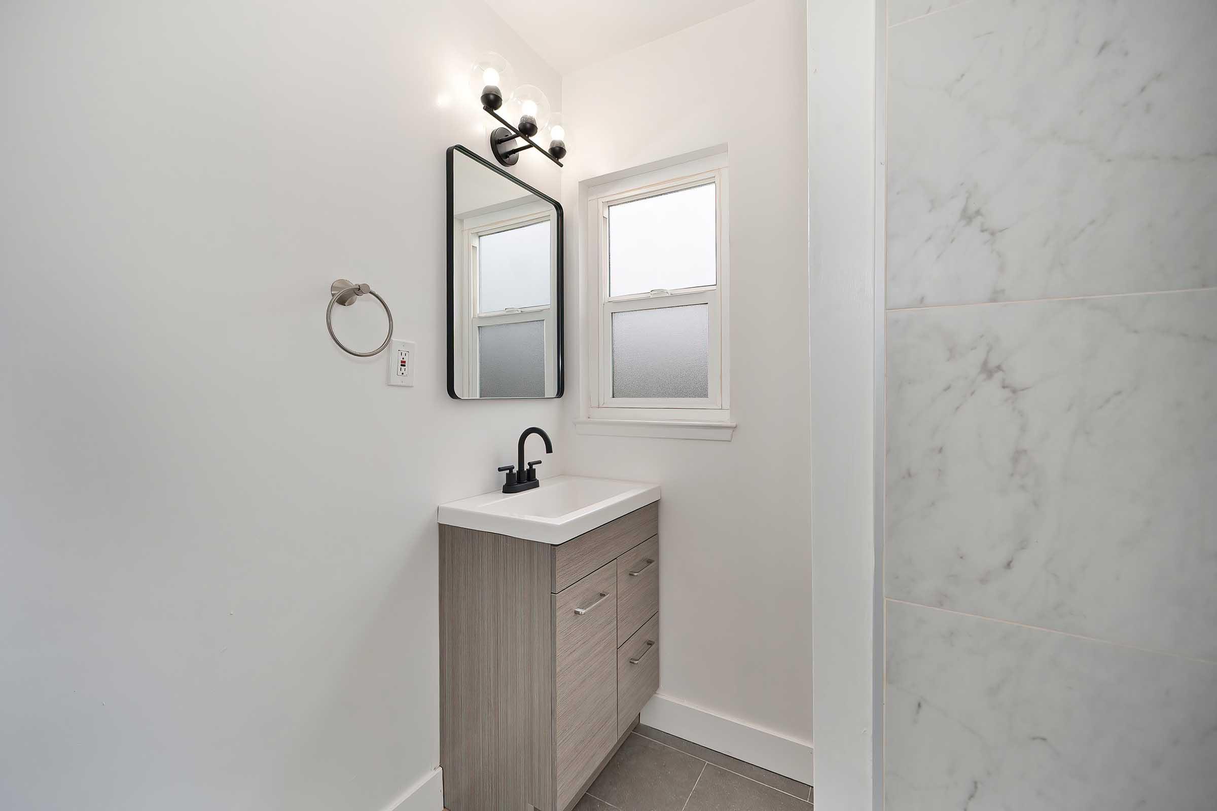 A modern bathroom featuring a light-colored wall, a minimalist vanity with a sink and wooden cabinetry, a large mirror above the sink, a wall-mounted light fixture, a small window, and a towel ring. The flooring is gray tile, and there is a marble-patterned wall section visible.