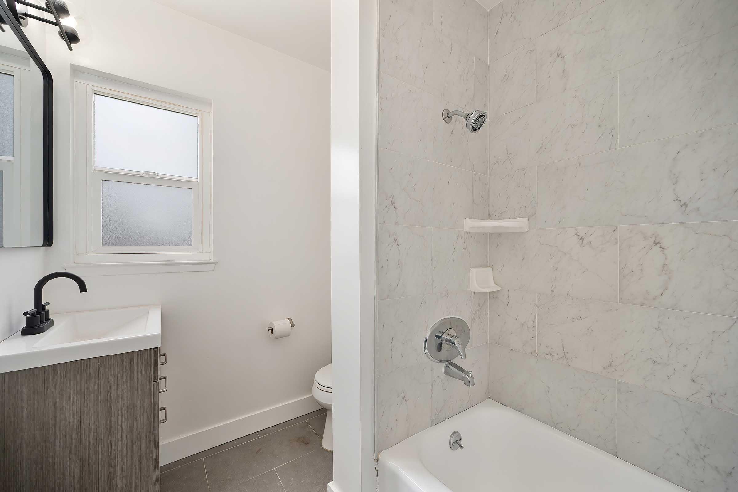 A modern bathroom featuring a white bathtub and shower with marble-style tiles, a sleek vanity with a black faucet, and a mirror. A small window allows natural light, and a toilet is visible in the background, along with a toilet paper holder on the wall.