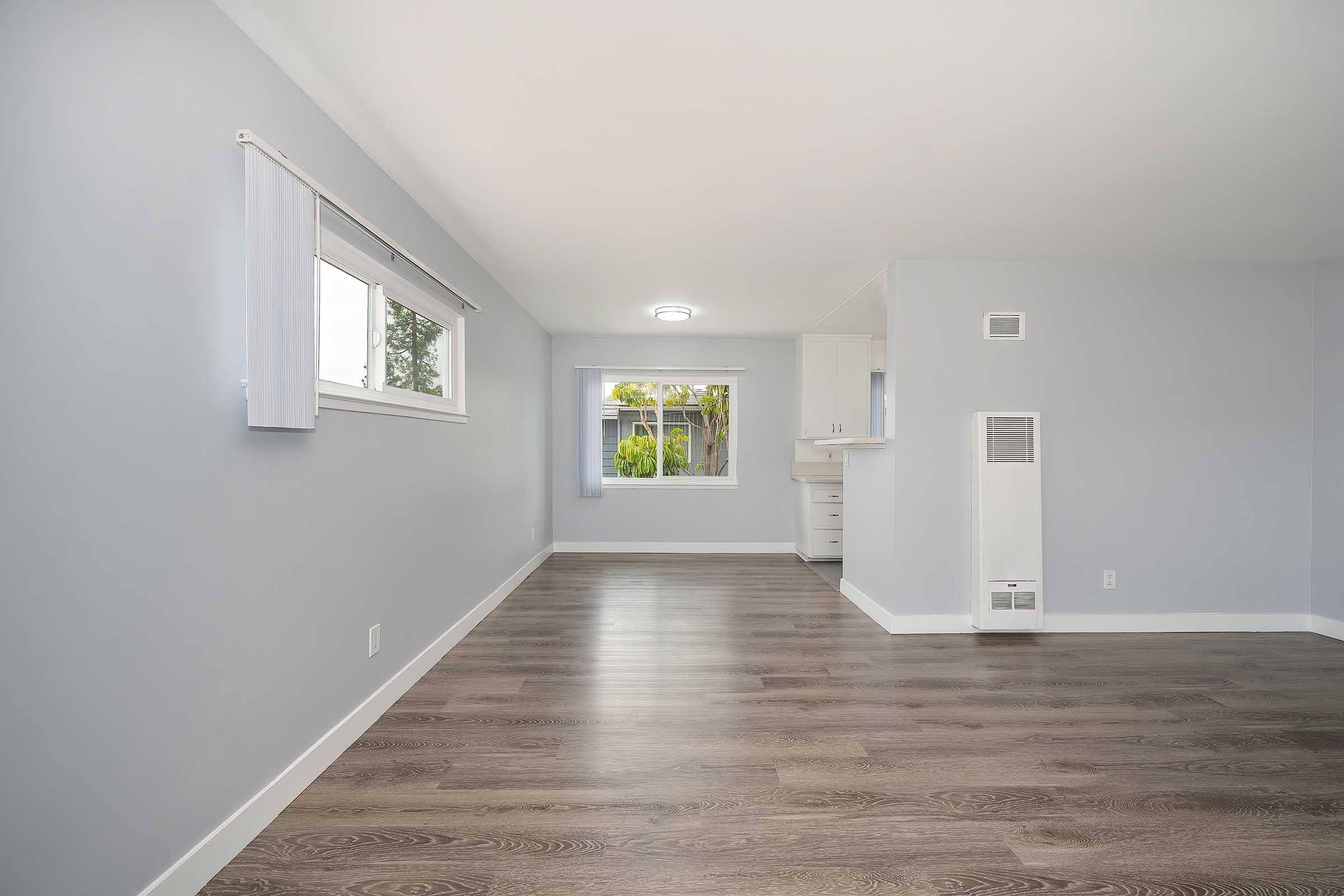 A spacious, bright room with light gray walls and hardwood flooring. There are large windows allowing natural light, a ceiling light fixture, and white window treatments. The room leads into a kitchenette area visible in the background, creating an inviting and open atmosphere.