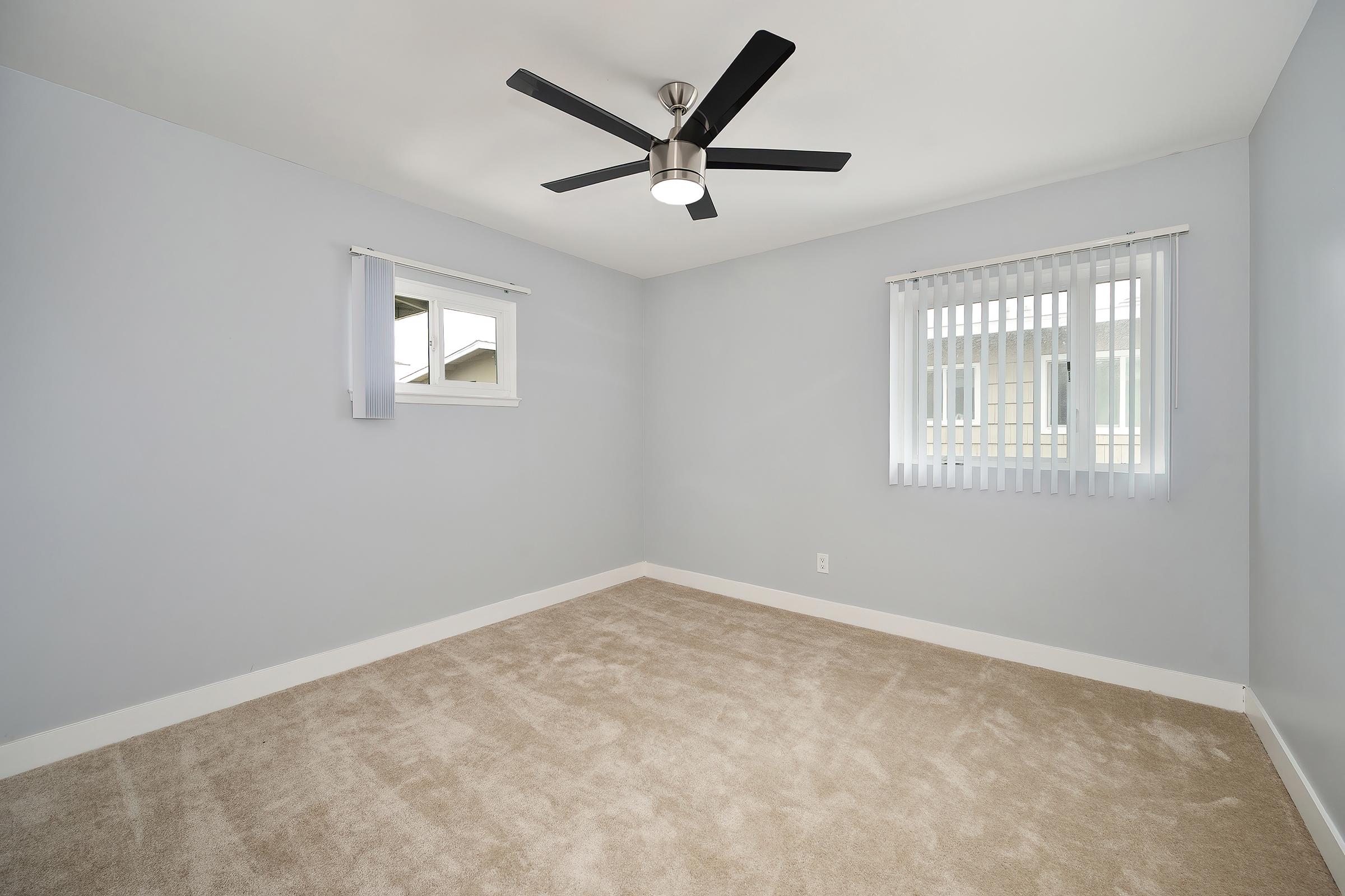 Empty room with light gray walls and a ceiling fan. The floor is carpeted in a light beige color. Two windows allow natural light, one with blinds, the other with curtains. The overall ambiance is clean and modern, creating a spacious feel.