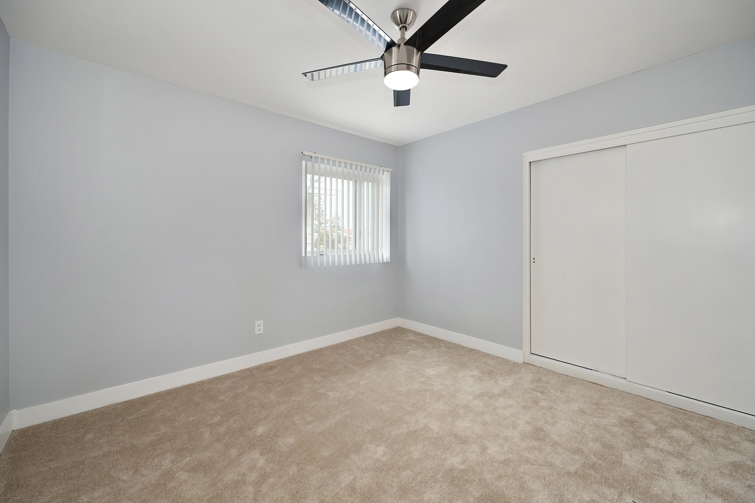 A simple and empty room featuring light gray walls, a ceiling fan with blades, and a window with horizontal blinds. The floor is covered in beige carpet, and there is a white closet with sliding doors on the right side of the image. The overall ambiance is clean and minimalistic.
