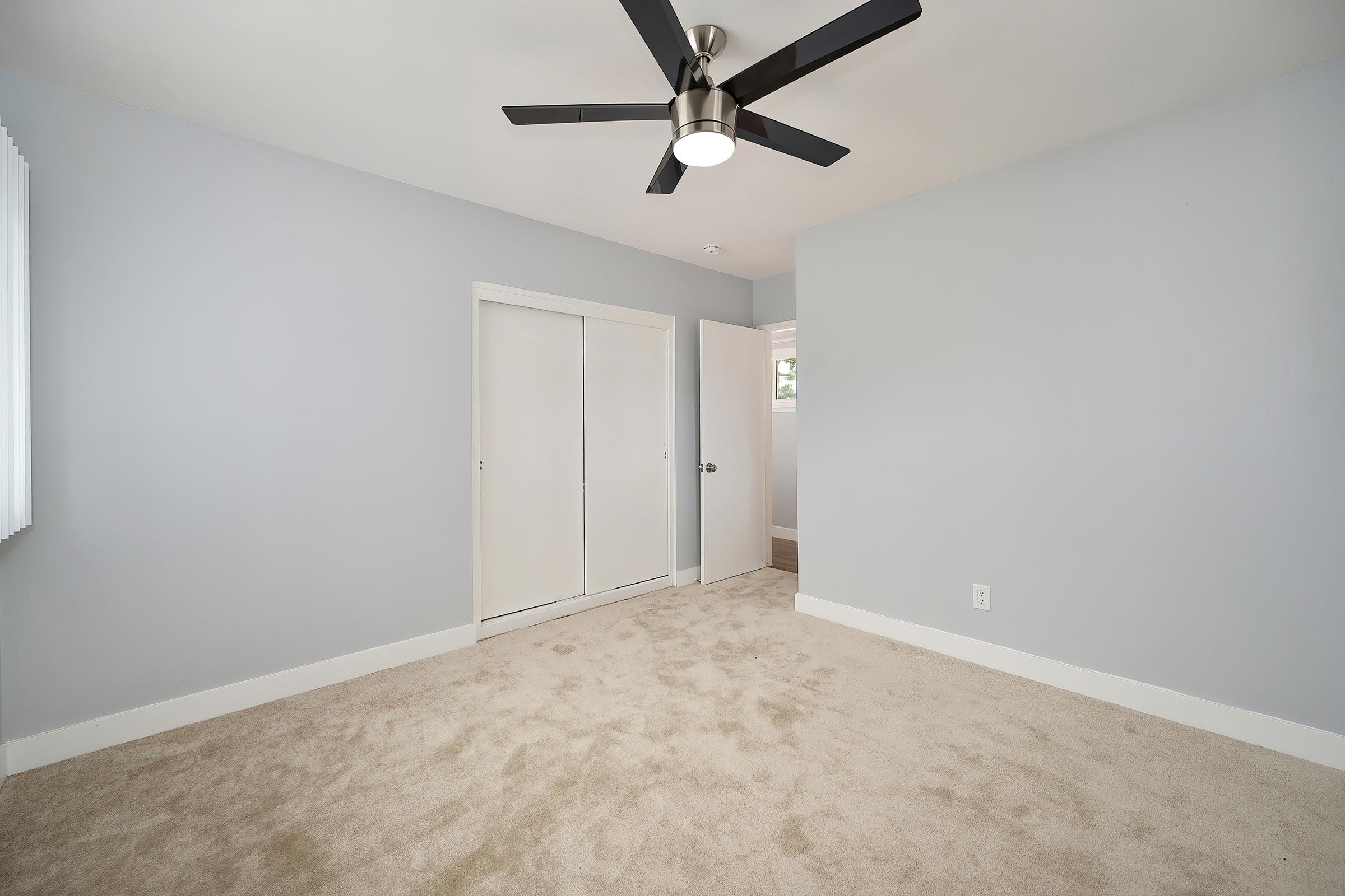 A minimalistic room featuring light gray walls, carpeted flooring, and a ceiling fan. There is a double closet with white sliding doors on one side and a doorway leading to another room or area. Natural light enters through a window, creating a bright and airy atmosphere.