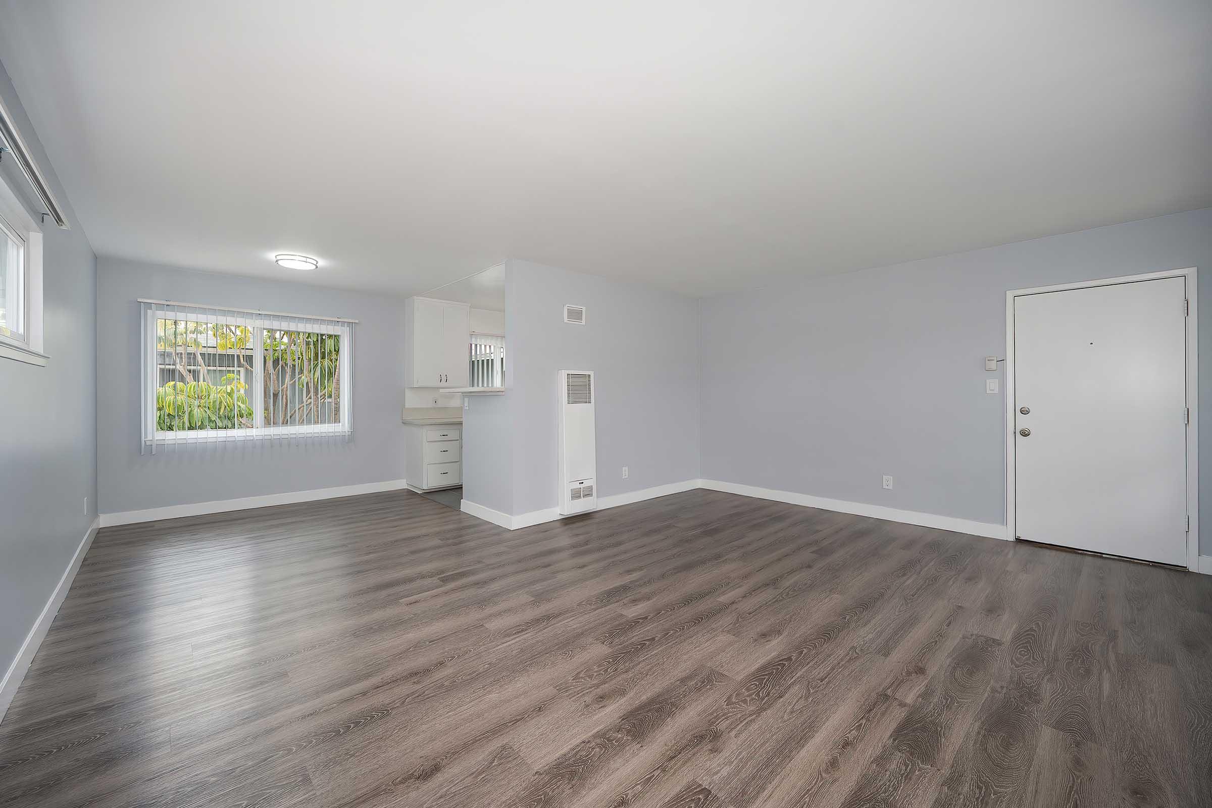 Spacious empty room with light gray walls and wood-like flooring, featuring a doorway on the right, large windows with blinds, and a small kitchen area visible in the background. Natural light fills the space, creating a bright and airy atmosphere.