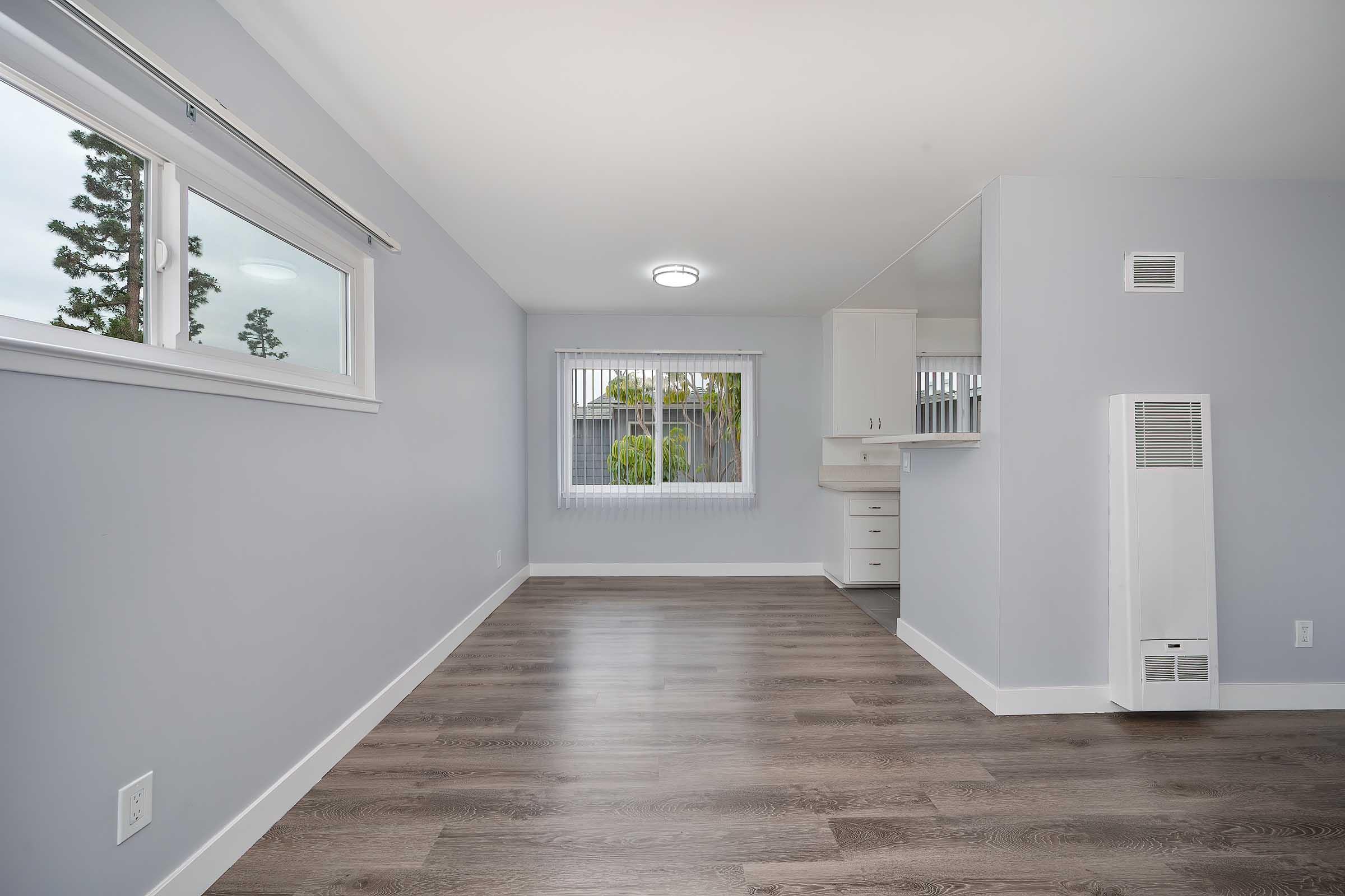 Bright and spacious room with light gray walls, large window with blinds, and modern wood flooring. The area features a ceiling light fixture and an open layout that connects to a kitchen or dining space, with a small wall-mounted heater on one side. Ideal for creating a cozy living environment.