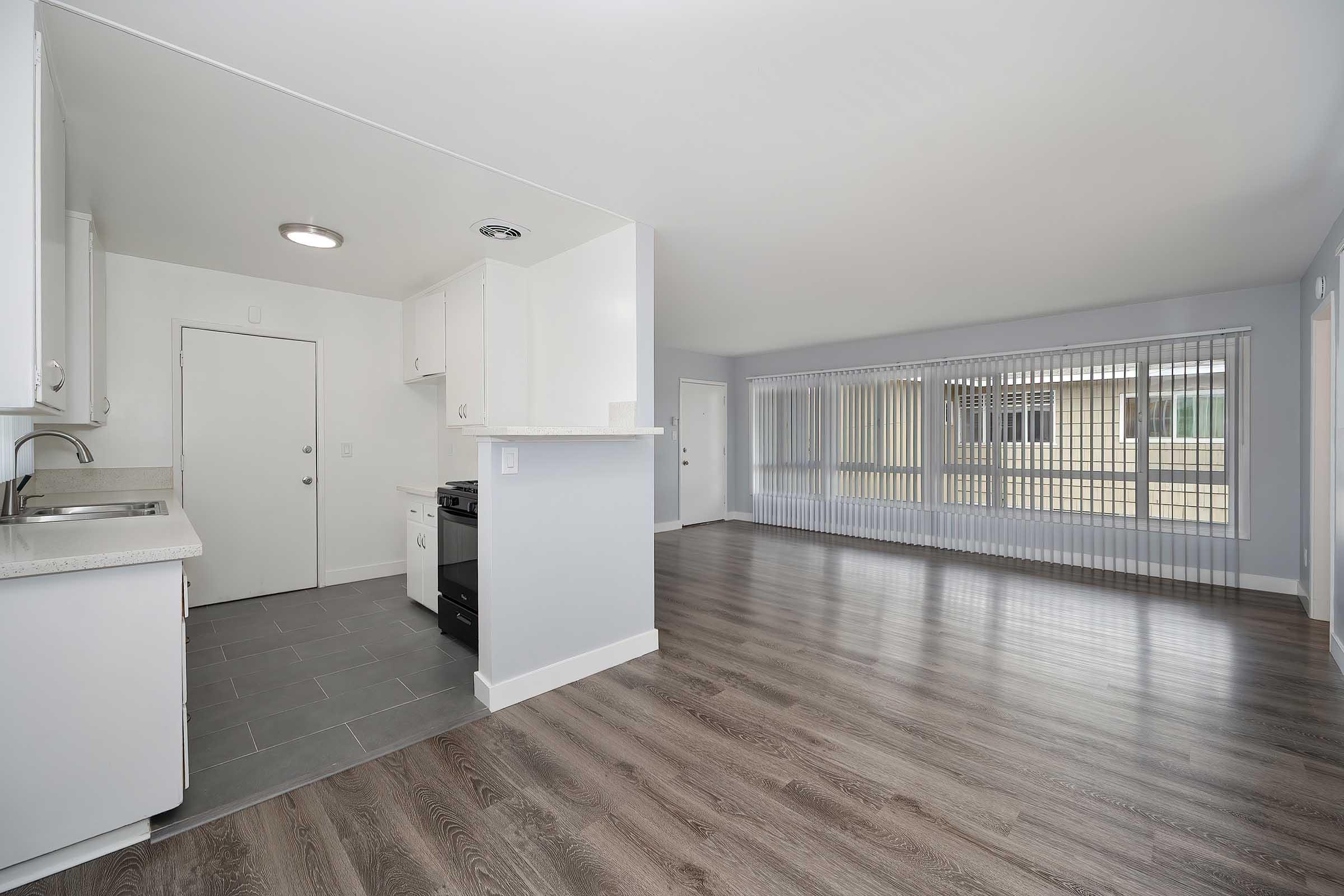 A modern kitchen area with white cabinets and a gas stove, featuring an open layout that connects to a spacious living area. Large windows with vertical blinds allow natural light to fill the room. The flooring is a sleek, light-colored laminate.