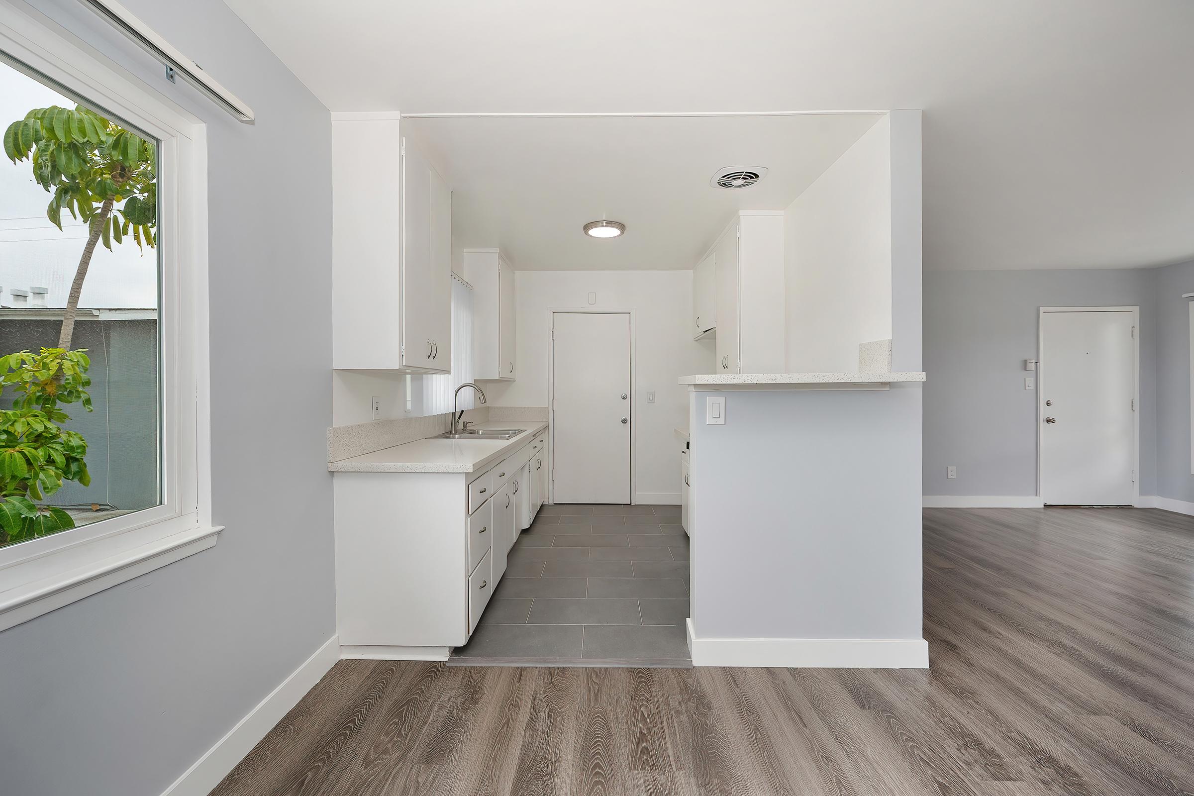 Modern kitchen and living area featuring white cabinetry, a solid surface countertop, and gray tiled flooring. The space is well-lit with natural light from a window, showcasing a clean and open layout. The walls are painted in a light gray hue, contributing to a contemporary aesthetic.