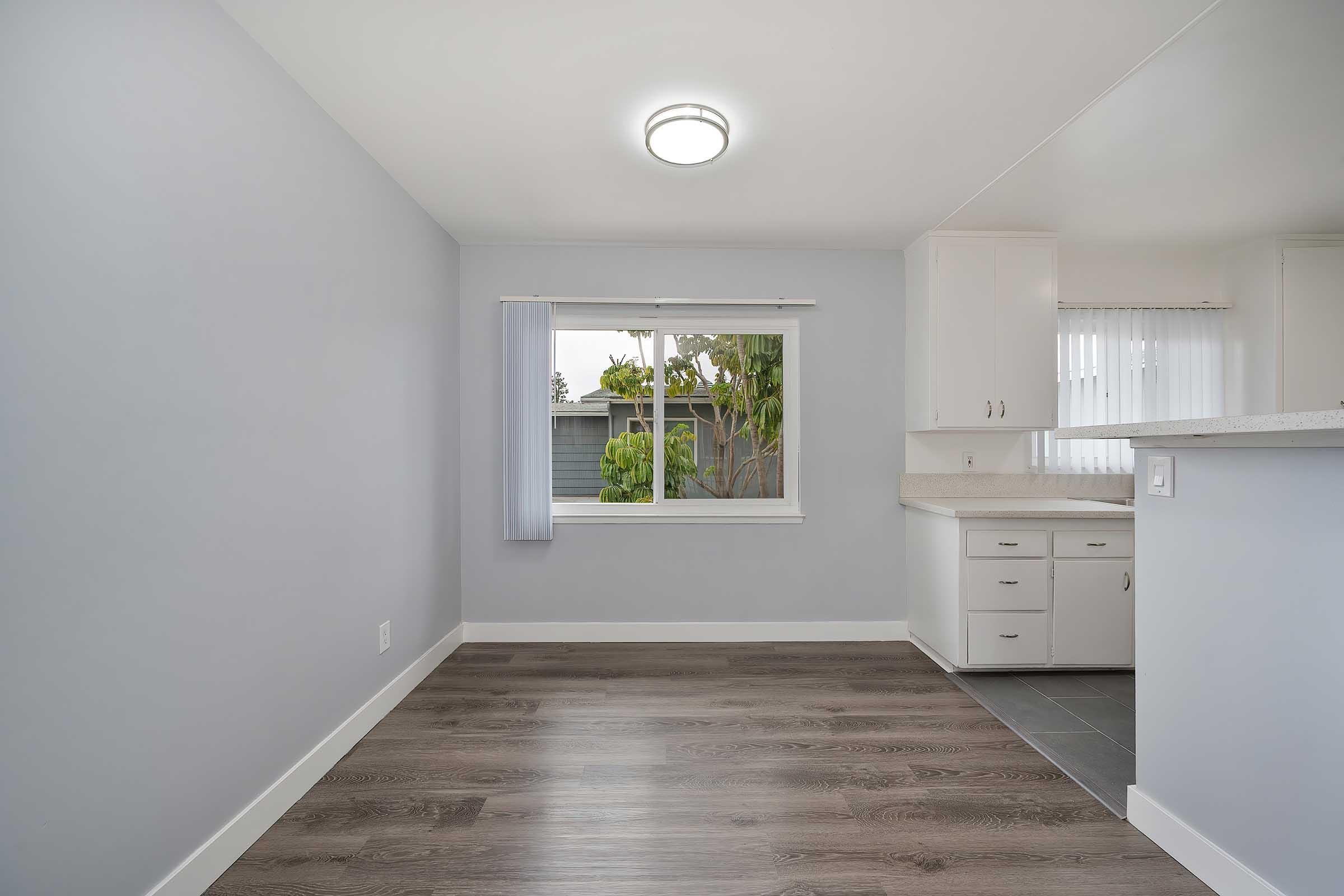 A brightly lit room with light gray walls and modern flooring. A window with white curtains offers views of greenery outside. The space features a small kitchen area with white cabinetry, creating a clean and open atmosphere, ideal for living or dining.