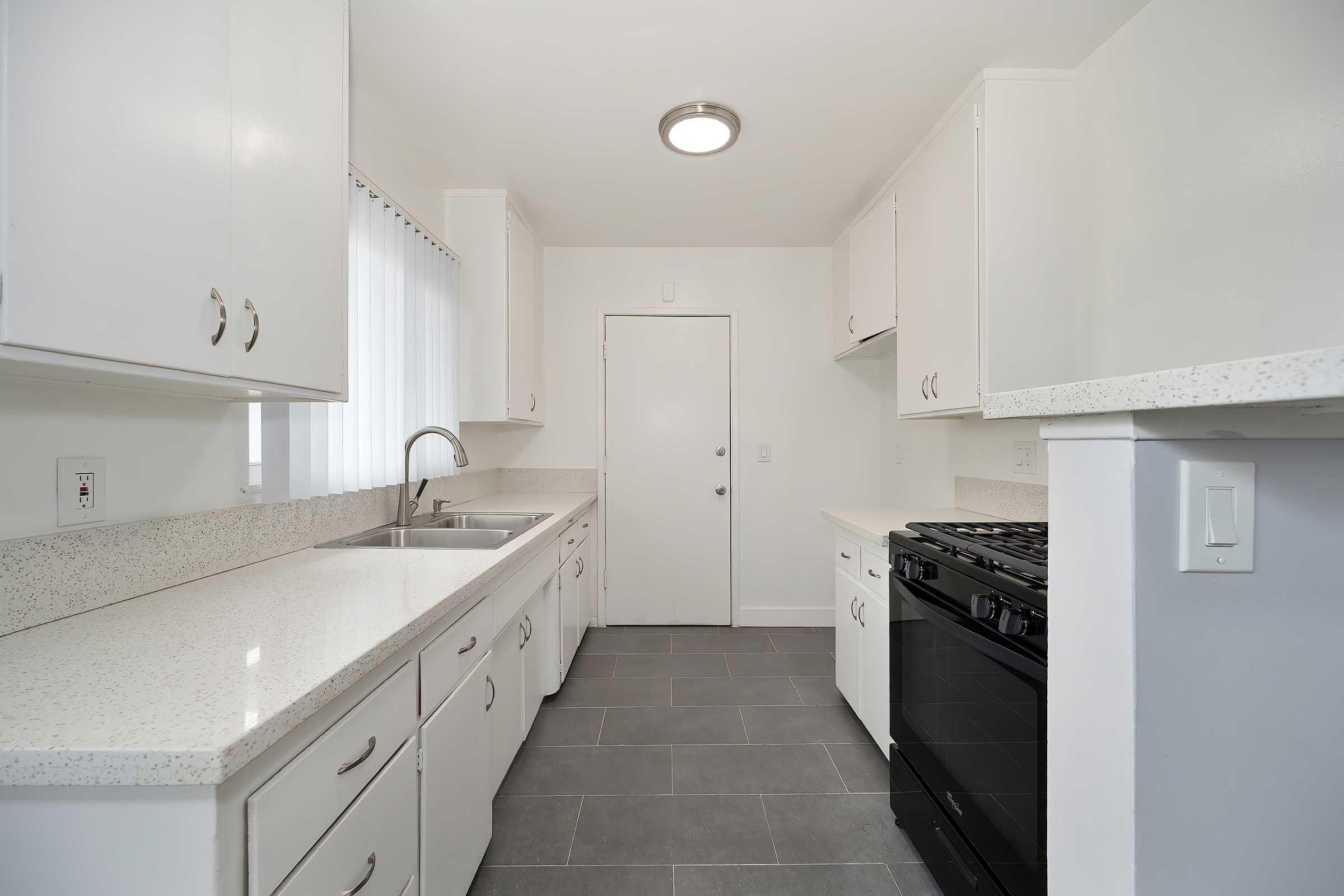A modern kitchen featuring white cabinetry, a countertop with speckled design, stainless steel sink, and black stove. Natural light comes in through a window, and a door leads to another room. The floor is tiled in grey, creating a clean and contemporary aesthetic.