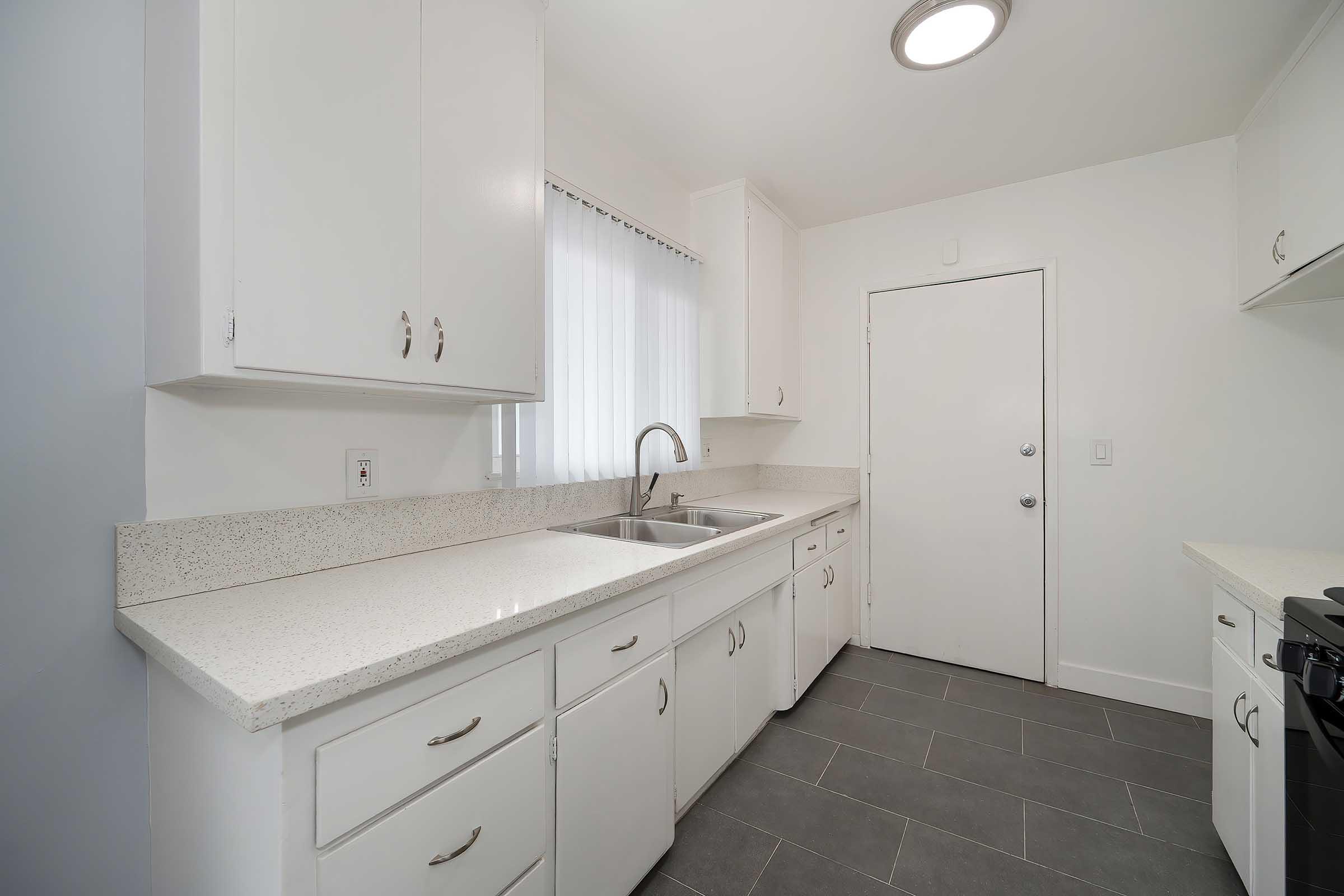 Modern kitchen with white cabinetry and a light-colored granite countertop. Features a double sink, a window allowing natural light, and a light fixture in the ceiling. The floor is tiled in a dark shade, and a door is visible at the far end of the kitchen.