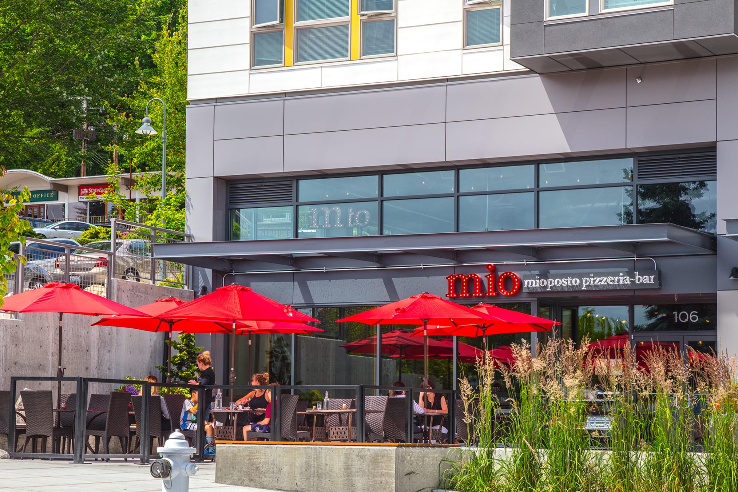 A modern pizzeria bar named "mio" with outdoor seating featuring red umbrellas.