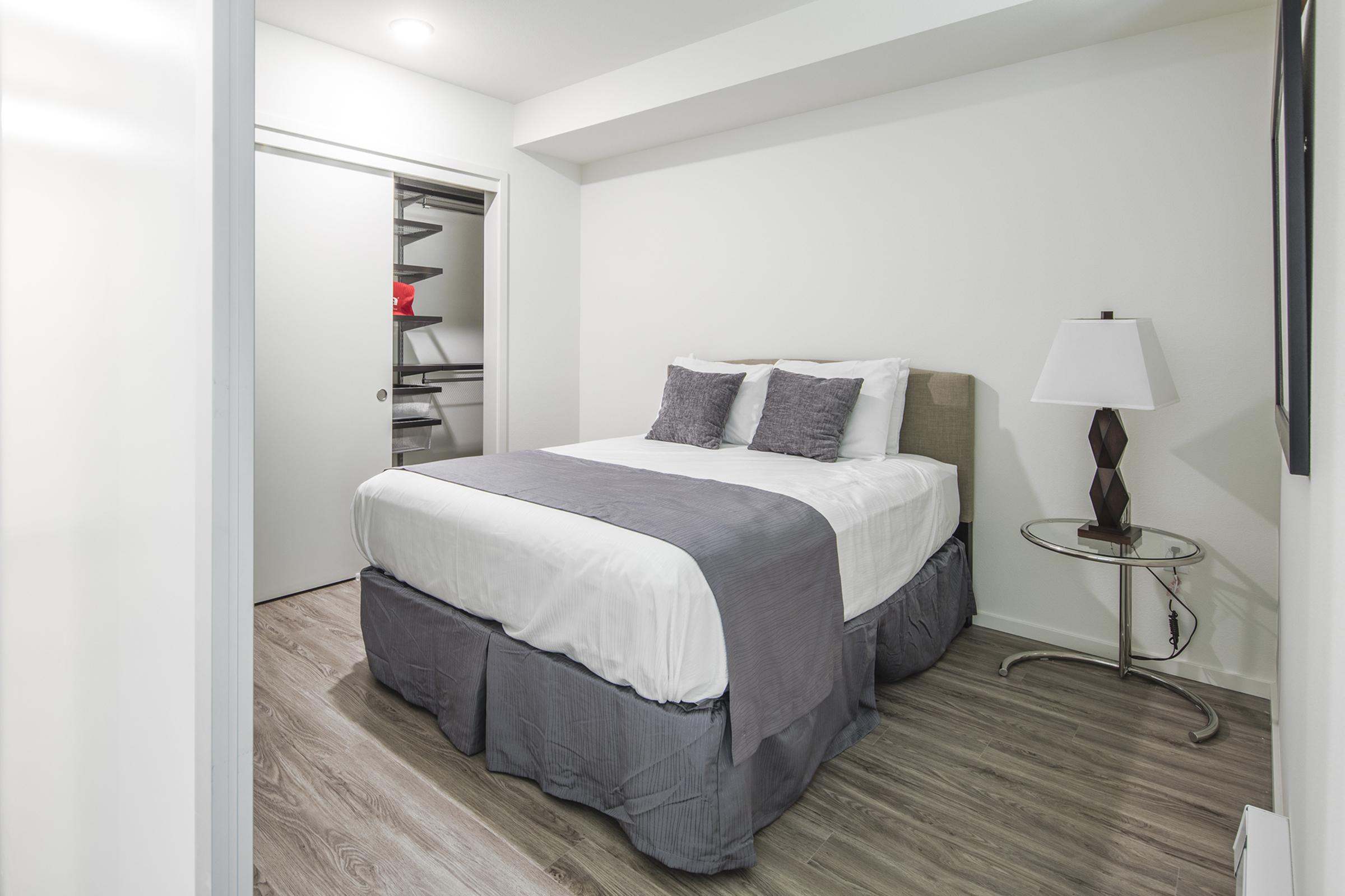 A cozy bedroom featuring a queen-sized bed with gray and white bedding, two decorative pillows, and a side table with a lamp. A closet is visible in the background, and the floor is covered with light-colored wood paneling. The walls are painted in a neutral tone, creating a calm ambiance.