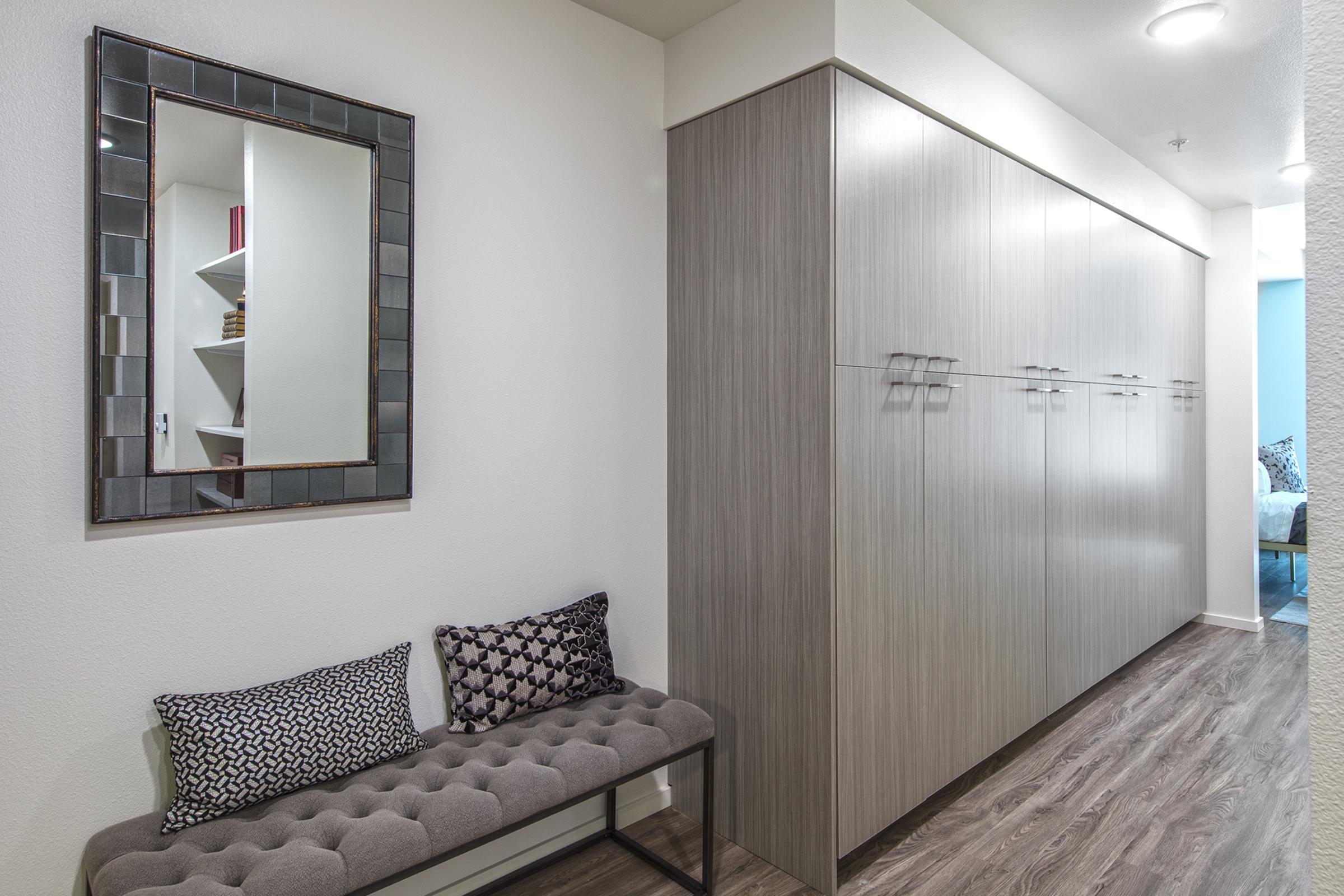 A modern hallway featuring light-colored walls and a large cabinet with multiple doors on one side. A stylish mirror with a decorative frame hangs above a tufted bench adorned with two patterned cushions. The floor is made of wood-like laminate, adding to the contemporary aesthetic.