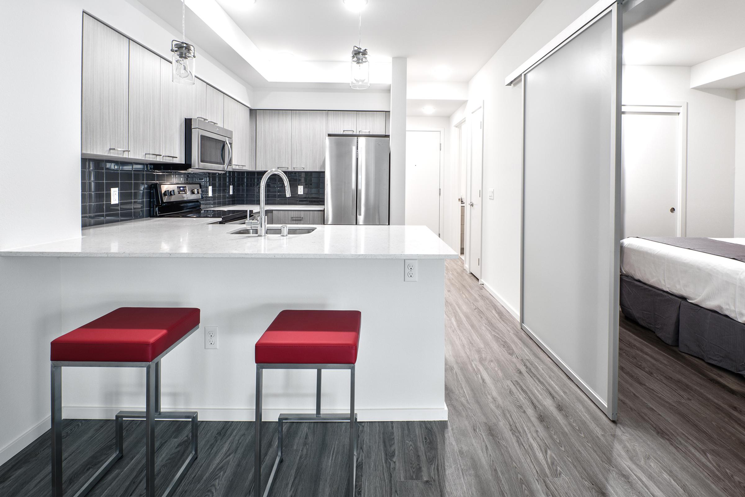 Modern kitchen with a sleek design, featuring a white countertop, stainless steel appliances including an oven and refrigerator, and a dark tile backsplash. Two red barstools are placed at the counter. A doorway leads to a bedroom area, and the flooring is a contemporary wood-like laminate.