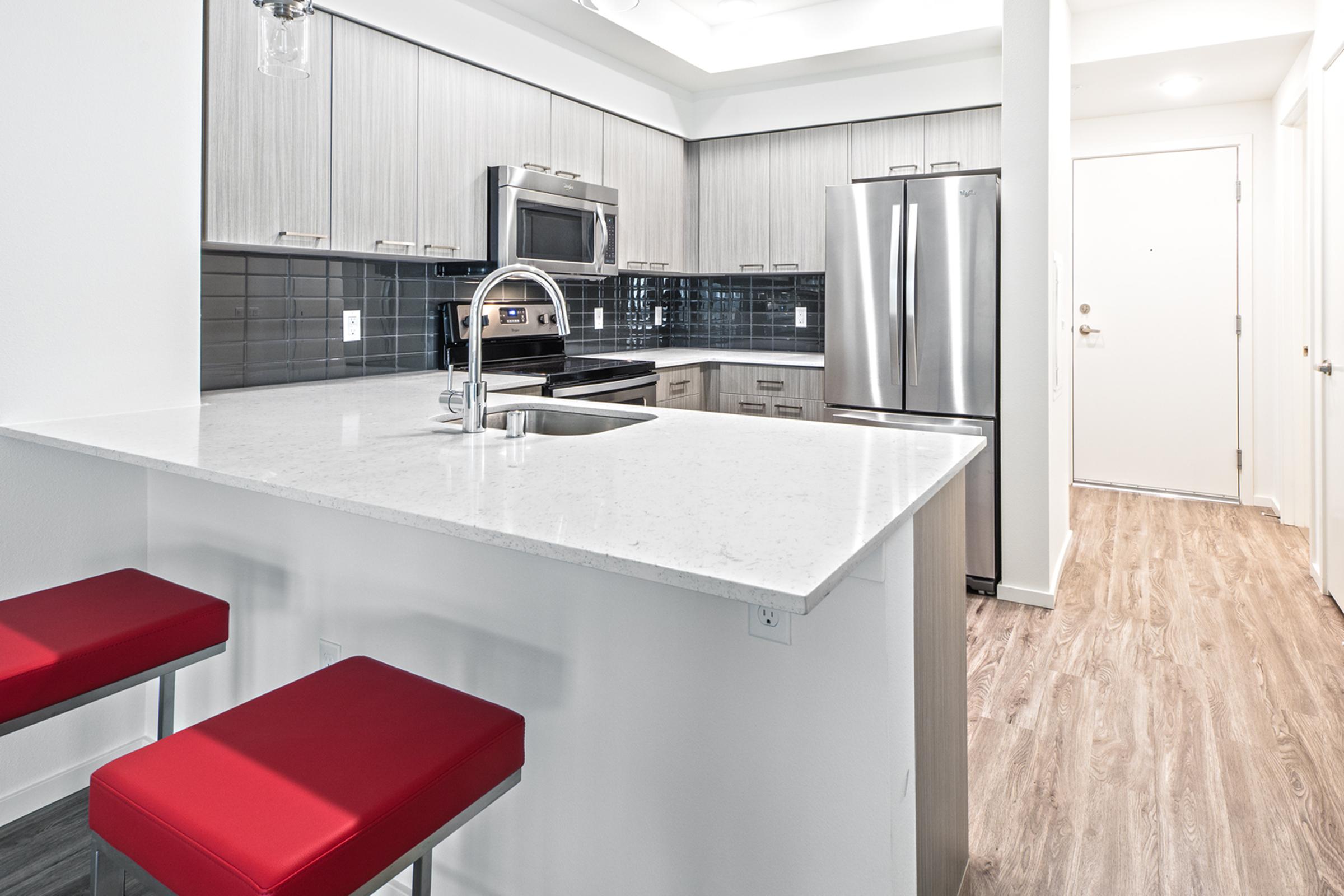 Modern kitchen featuring a sleek design with grey cabinets, stainless steel appliances, a white countertop, and a breakfast bar with red stools. The backsplash is dark blue tiles, adding contrast, while the flooring is light wood. Natural light illuminates the space, making it inviting and contemporary.