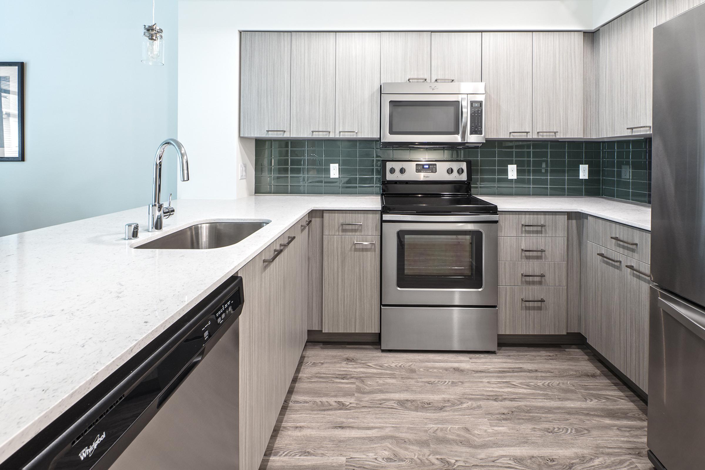 Modern kitchen featuring light wood cabinetry, a stainless steel oven and microwave, a sleek sink with a faucet, and a green tile backsplash. The countertop is a light-colored stone, and the kitchen has an open layout with integrated appliances, including a dishwasher. Hardwood flooring completes the contemporary look.