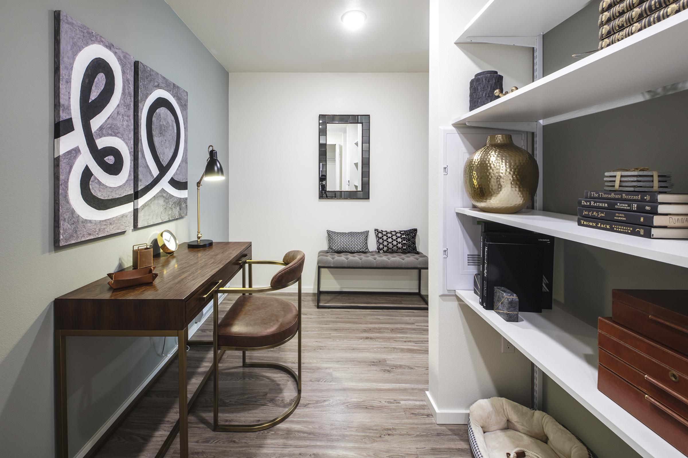 A modern home office featuring a sleek wooden desk with a lamp, a decorative mirror on the wall, and abstract artwork. There is a padded bench against one wall and a white shelf unit displaying books and decorative items. The room has neutral colors and warm lighting, creating a cozy ambiance.
