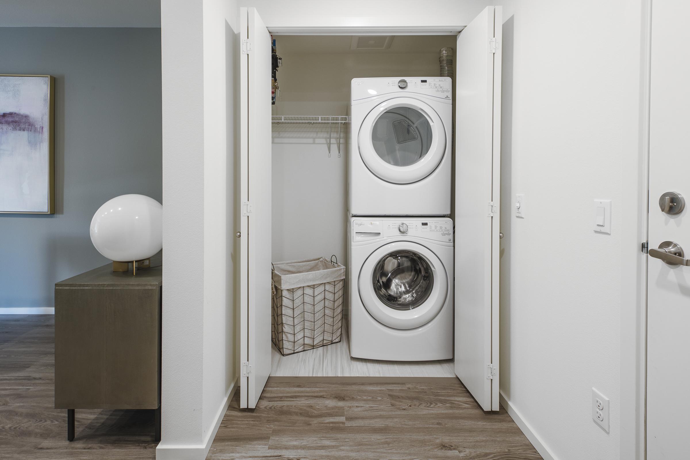 A laundry nook featuring a stacked washer and dryer in a clean, well-lit space. There's a decorative lamp on a side table and a wicker basket for laundry, with closet doors slightly ajar. The walls are painted a soft color, enhancing the modern, minimalistic aesthetic.