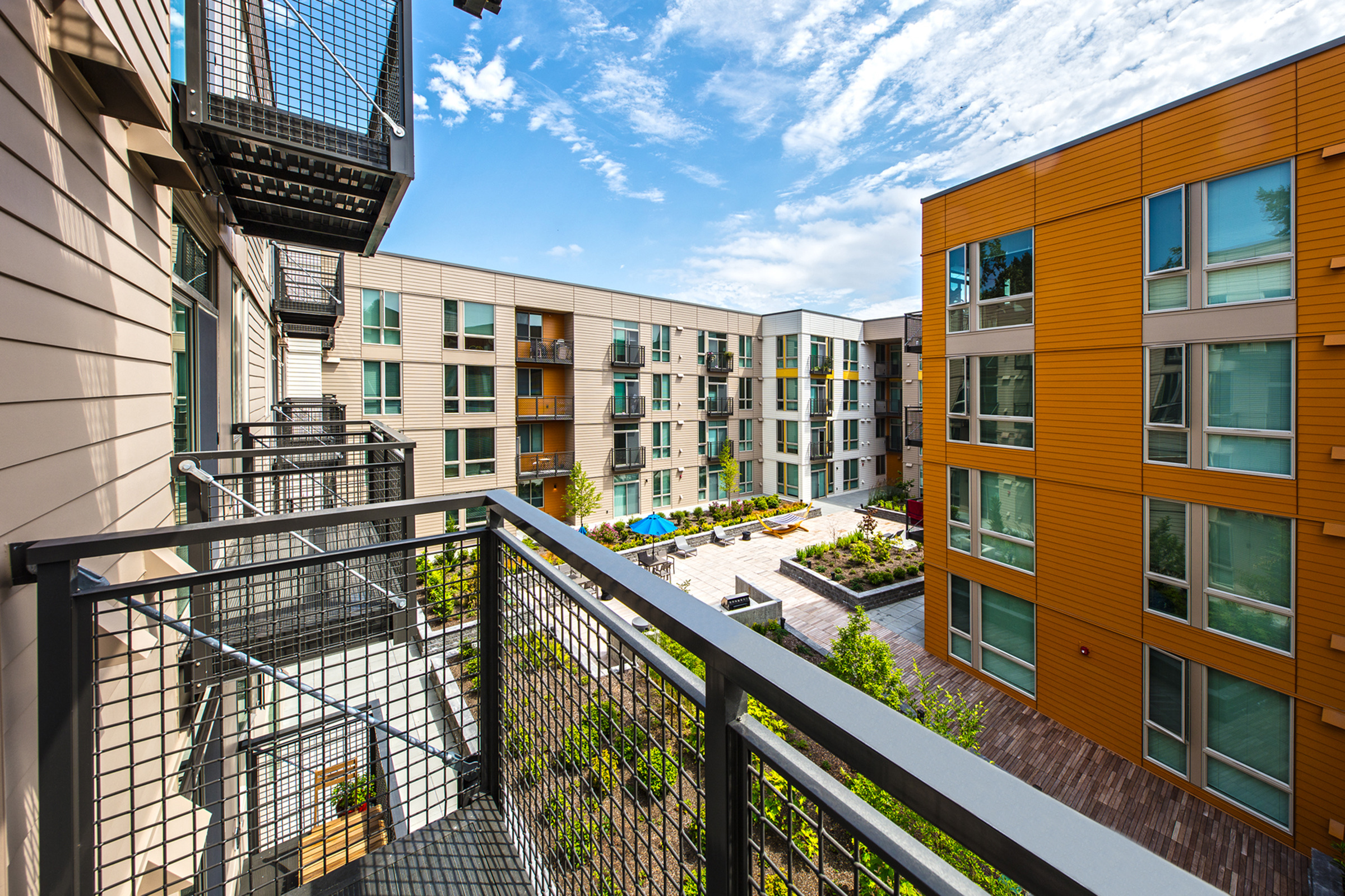 View from a balcony overlooking a modern apartment complex. The scene features two buildings with varying architectural styles, surrounded by greenery and a landscaped courtyard. A blue pool is visible in the distance under a bright sky with scattered clouds.