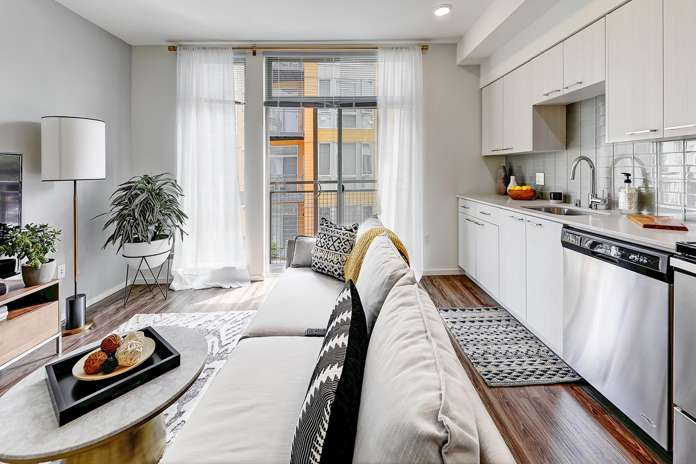 Bright and modern living space featuring a cozy sofa with decorative pillows, a coffee table with snacks, and a sleek kitchen area with white cabinets and stainless steel appliances. Large windows allow natural light to fill the room, complemented by indoor plants and stylish decor.