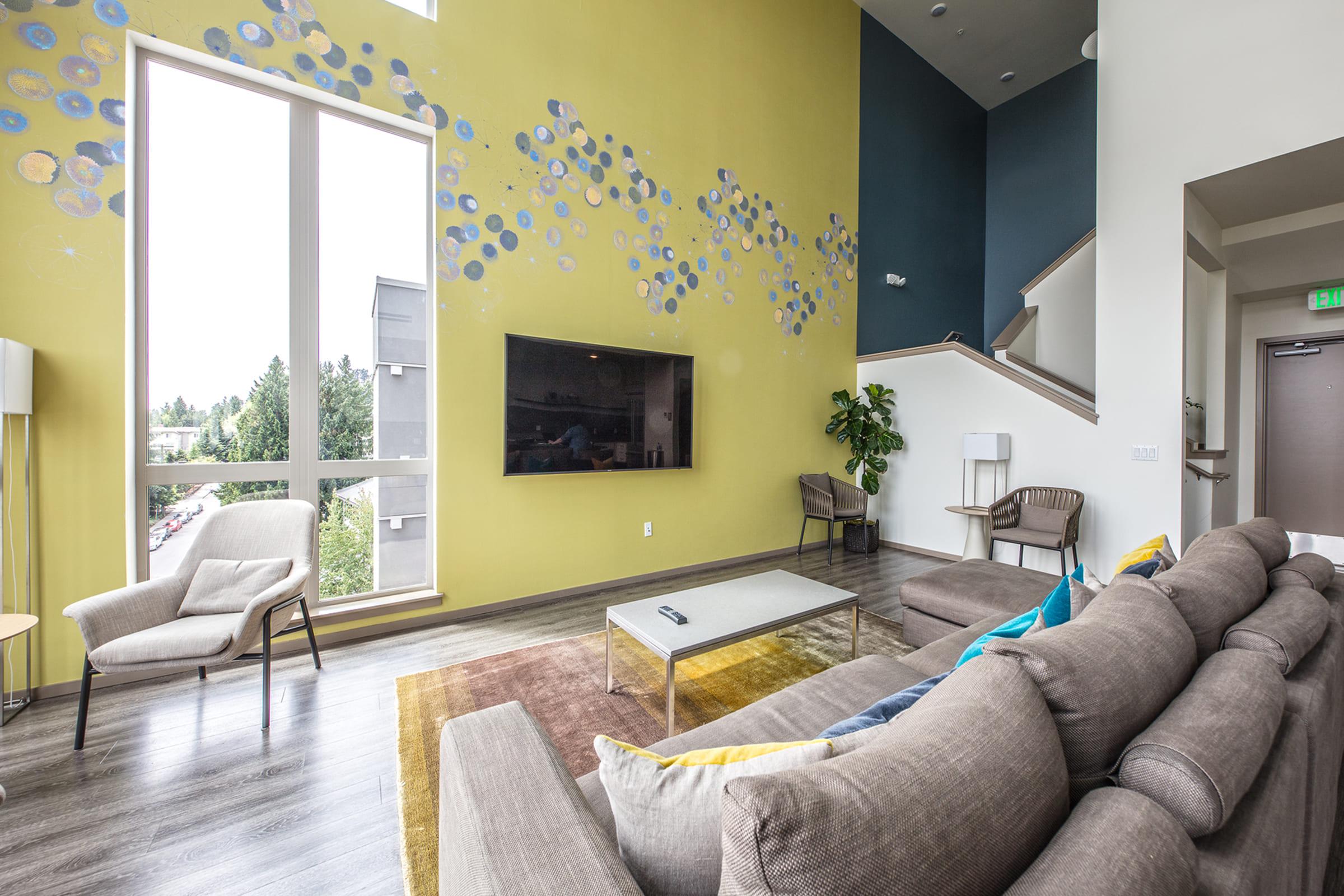 A modern living room featuring a large window with natural light, a gray sectional sofa, a coffee table, and a wall-mounted TV. The room has vibrant yellow and blue wall artwork and a potted plant in the corner, creating a stylish and inviting atmosphere.