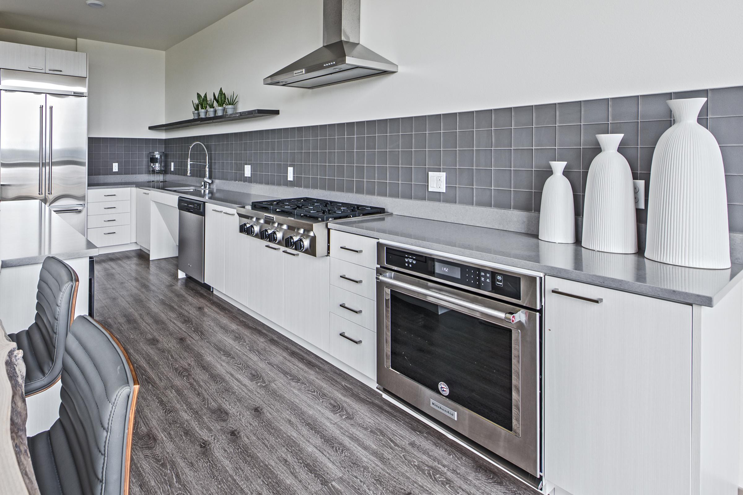 Modern kitchen featuring sleek cabinetry, stainless steel appliances, and a gray tile backsplash. A large gas stove and oven are prominently displayed, along with stylish white vases. The flooring is a warm wood tone, and the space is well-lit, creating a contemporary and inviting atmosphere.
