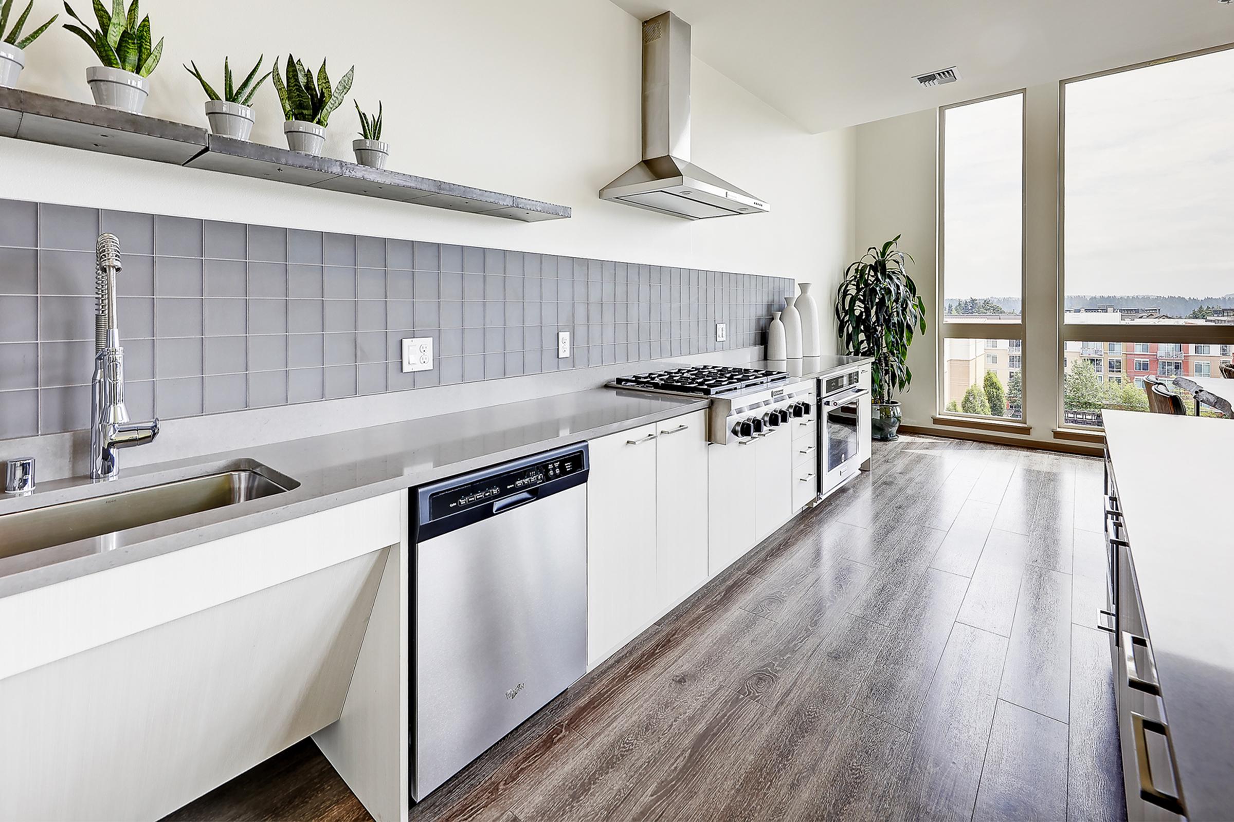 Modern kitchen featuring a sleek design with gray tiled backsplash, stainless steel appliances including a dishwasher and gas stove, and light wood flooring. Large windows allow natural light to flood the space, complemented by potted plants on the countertop for a touch of greenery.
