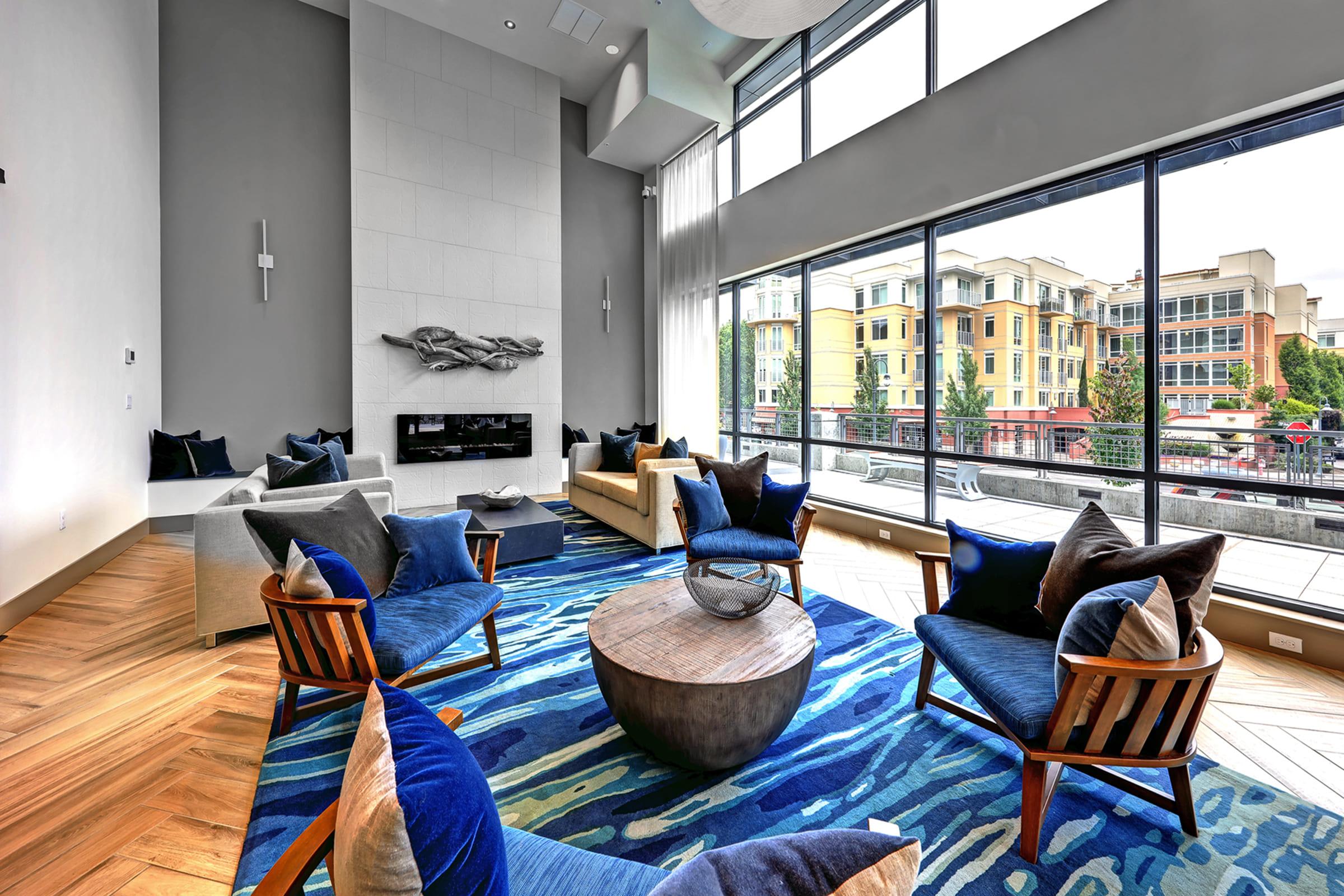 A modern lounge area featuring large windows with a view of an urban setting. The room is furnished with comfortable seating, including plush armchairs and sofas, arranged around a round coffee table. A stylish area rug adds color, and a contemporary fireplace is positioned on the wall.