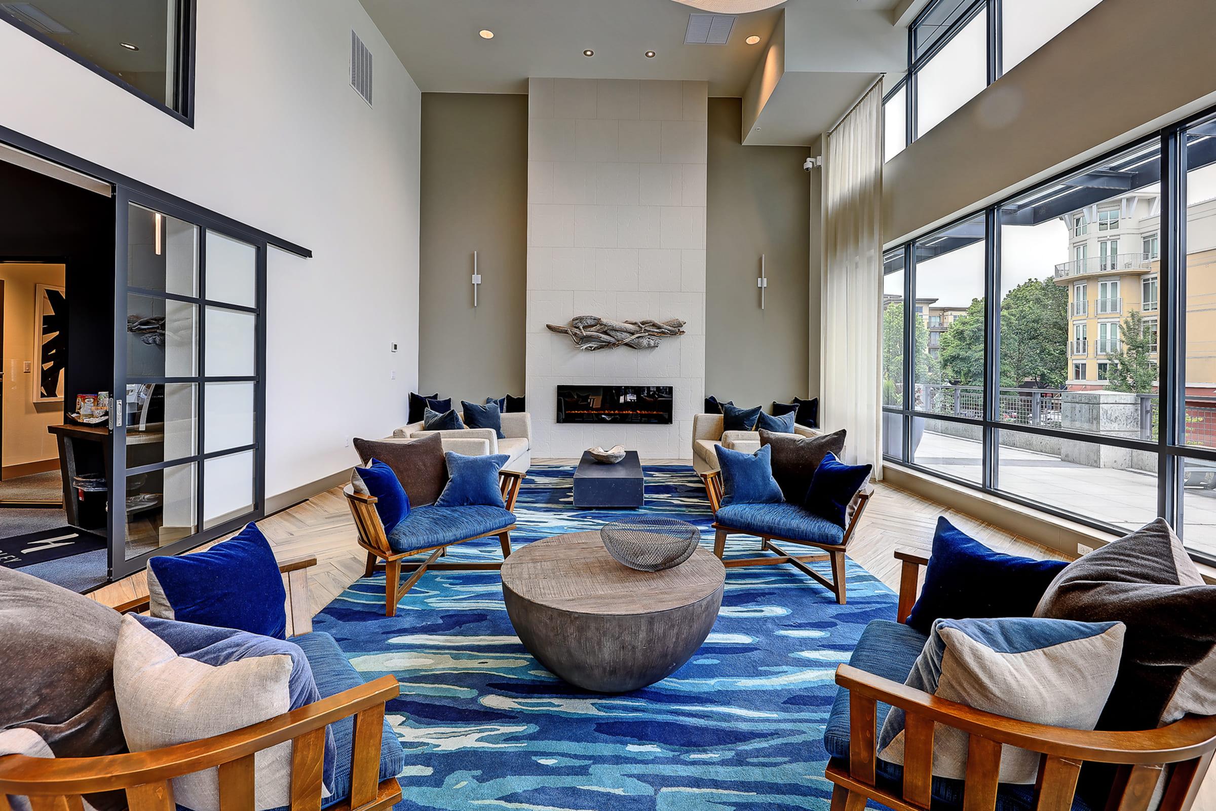 A modern lounge area featuring a fireplace, surrounded by comfortable seating with blue and beige cushions. The room has large windows allowing natural light, and a patterned blue rug complements the decor. The walls are neutral in color, creating a welcoming atmosphere.