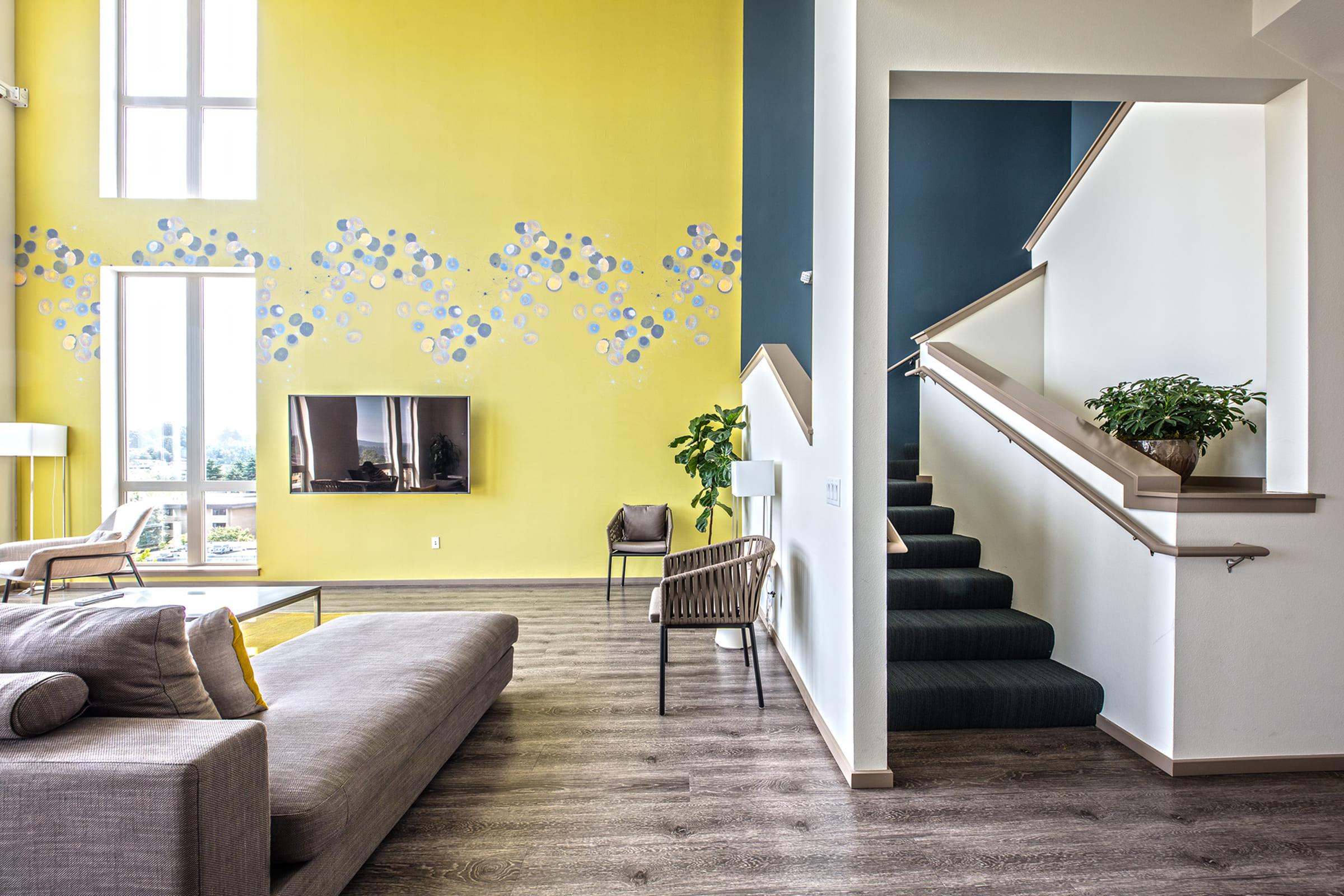 Bright and modern living space featuring a yellow accent wall with blue circular designs. A large window allows natural light, illuminating a gray sofa, minimalist decor, and a television mounted on the wall. Stairs lead to an upper level, with a green potted plant adding a touch of nature.