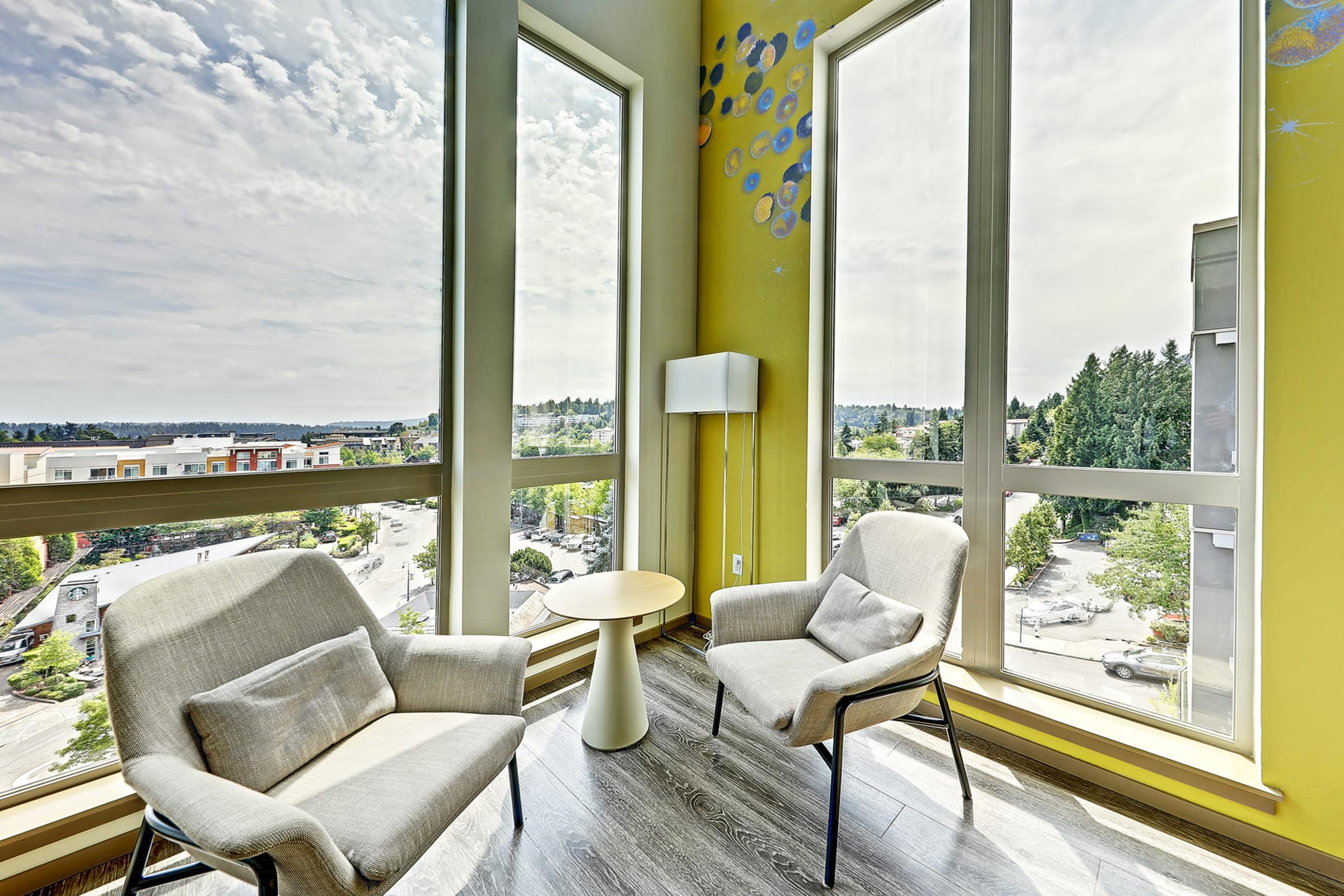 A bright and modern corner seating area with two light-colored chairs and a small round table. Large windows provide a panoramic view of an urban landscape and blue skies, while a white lamp stands nearby. The walls are painted a soft yellow, adding warmth to the space.