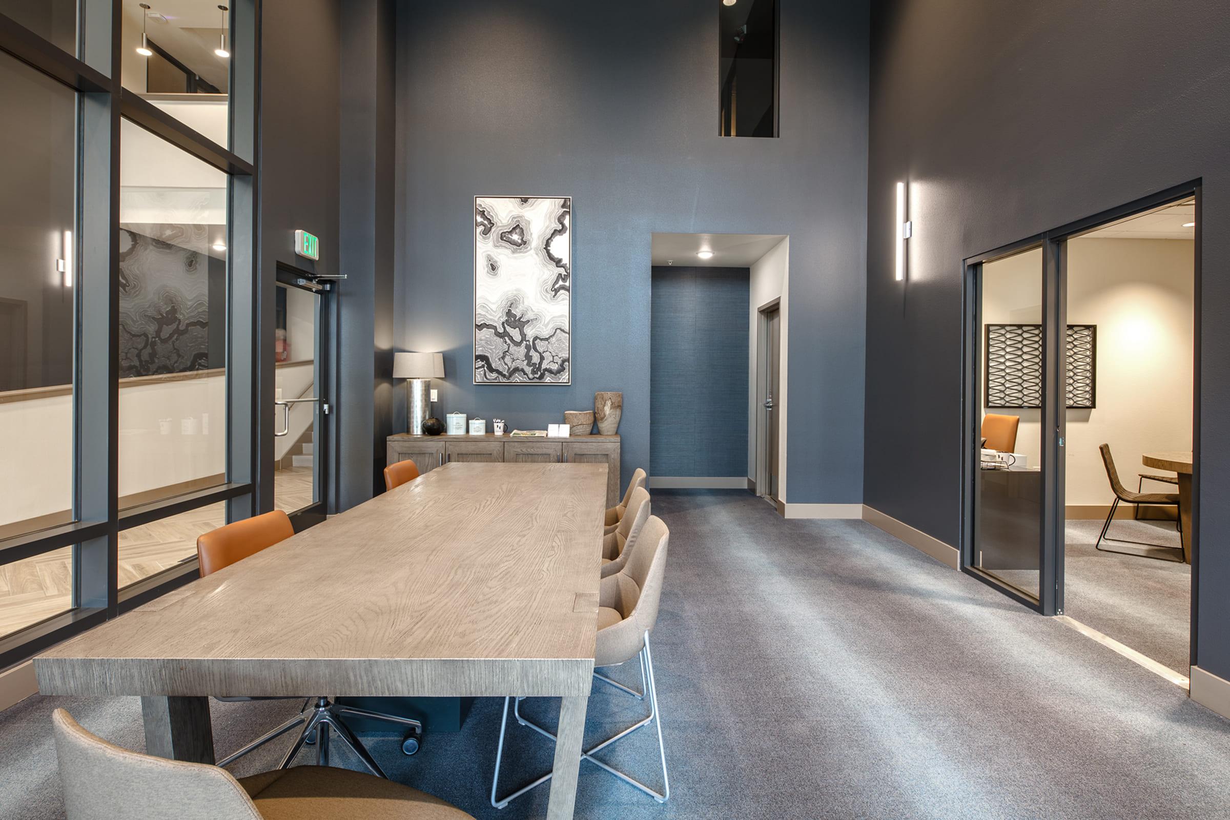 A modern conference room featuring a large wooden table surrounded by stylish chairs. The walls are painted in dark tones, with a large abstract painting on one wall. Glass doors lead to other office spaces, and soft lighting complements the contemporary decor. The floor has a sleek carpet design.