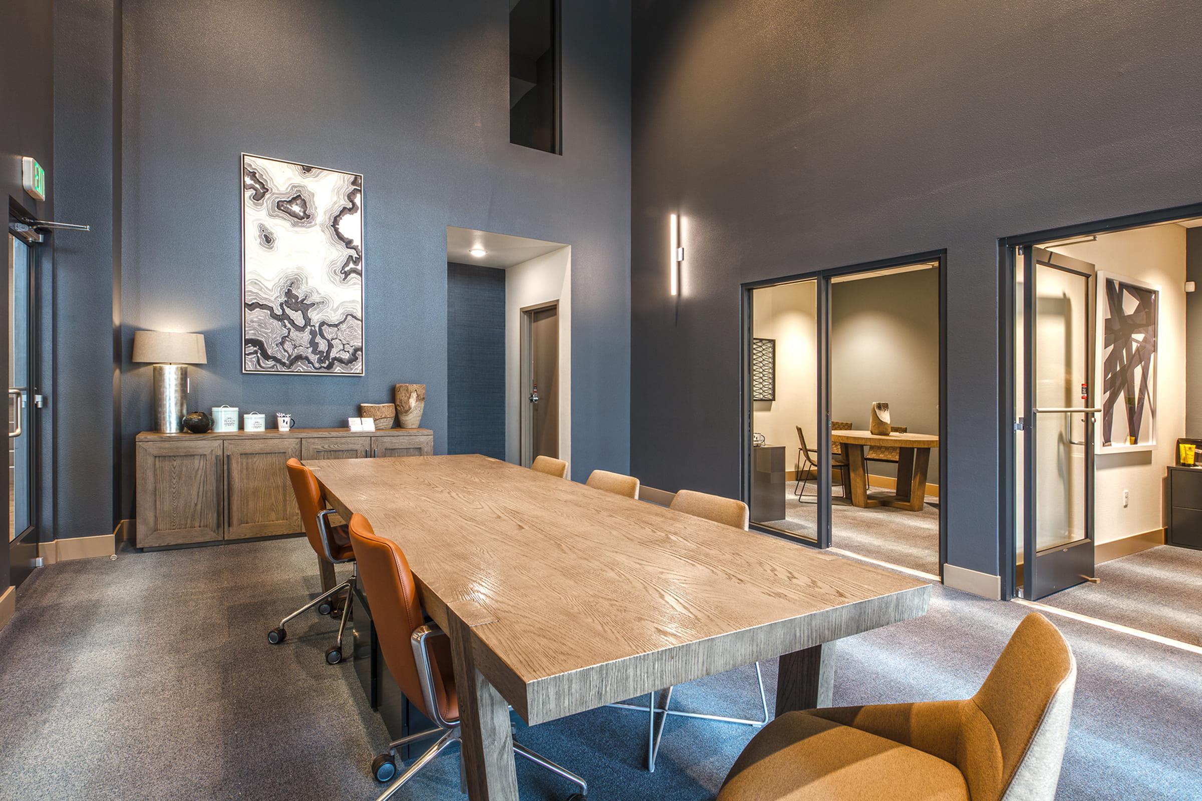 A modern office conference room featuring a large wooden table with rolling chairs, styled in neutral tones. The walls are dark with abstract artwork. Two glass-doored offices are visible in the background, along with a decorative lamp and a sideboard. Bright natural light enters through a window.