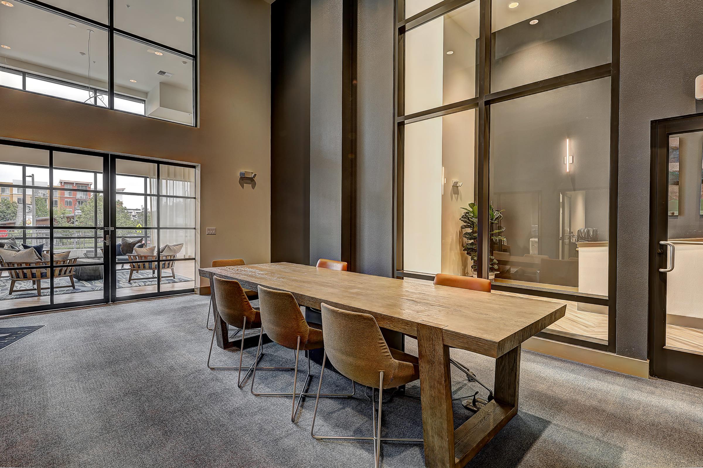 A modern conference room featuring a large wooden table surrounded by six chairs. The room has large windows allowing natural light, showcasing a view of an outdoor seating area. The decor is sleek and contemporary, with neutral tones and a plant in the corner.