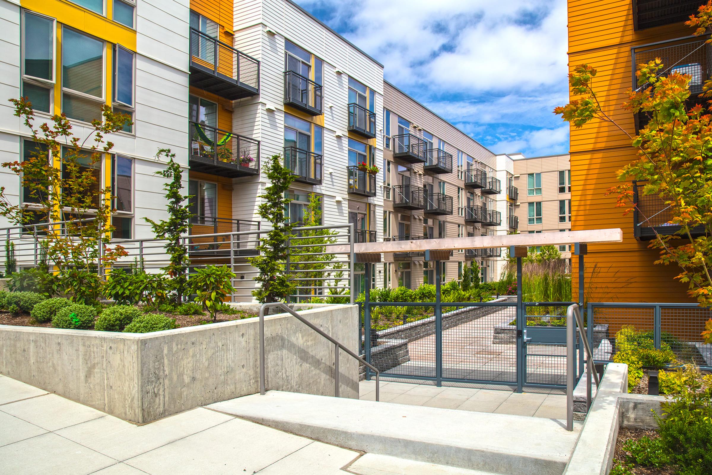 Modern apartment complex featuring two distinct buildings with various balconies. In the foreground, a landscaped area with low-maintenance plants and a concrete pathway leads to a gated entry, enhancing accessibility and inviting outdoor space. The backdrop includes a blue sky with scattered clouds.