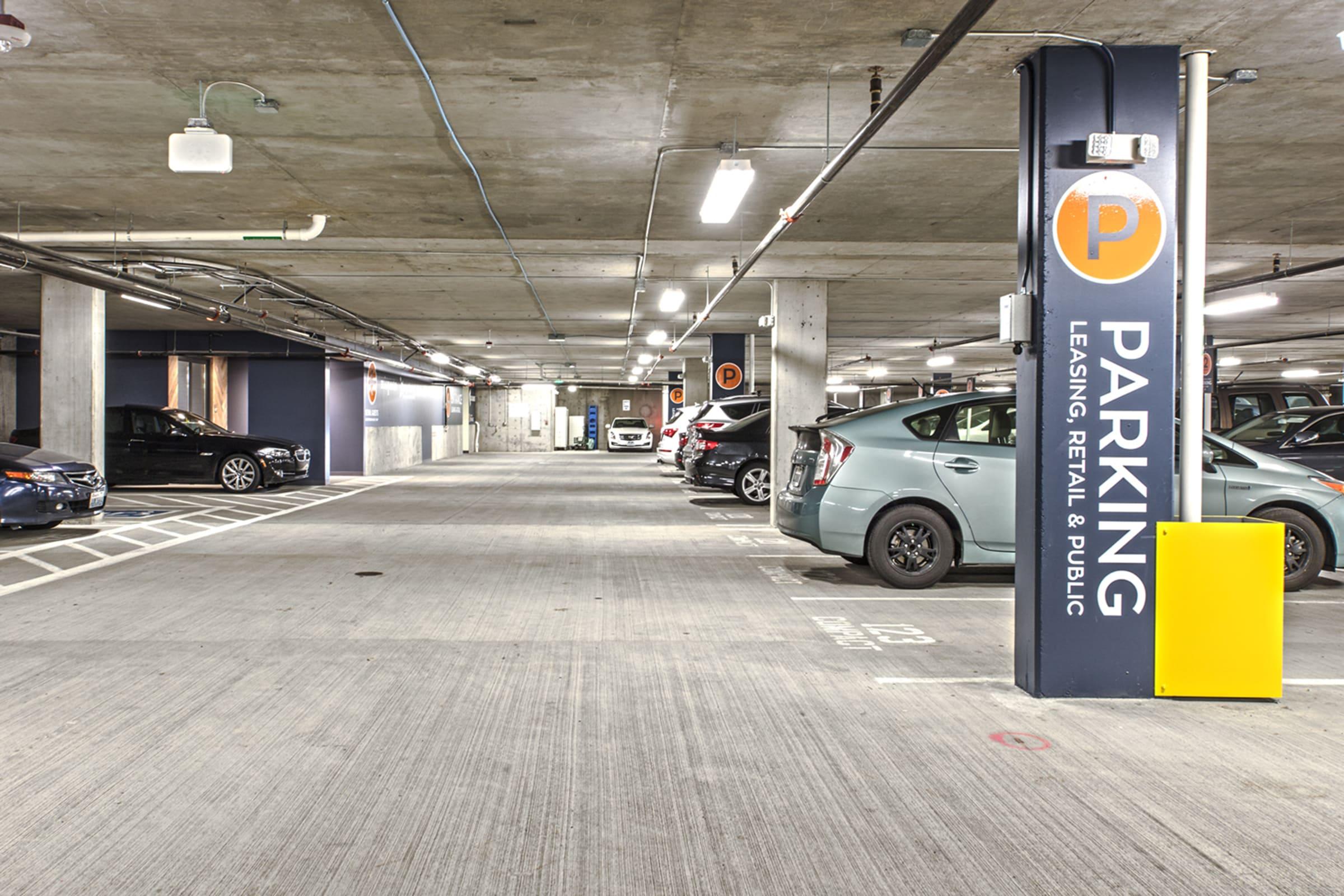 A spacious underground parking garage with empty parking spaces, well-lit by ceiling lights. There are several parked cars, including a grey car and a blue car. A sign indicating "PARKING: LEASING, RETAIL & PUBLIC" is prominently displayed on a pillar. The concrete floor is clean and clearly marked.