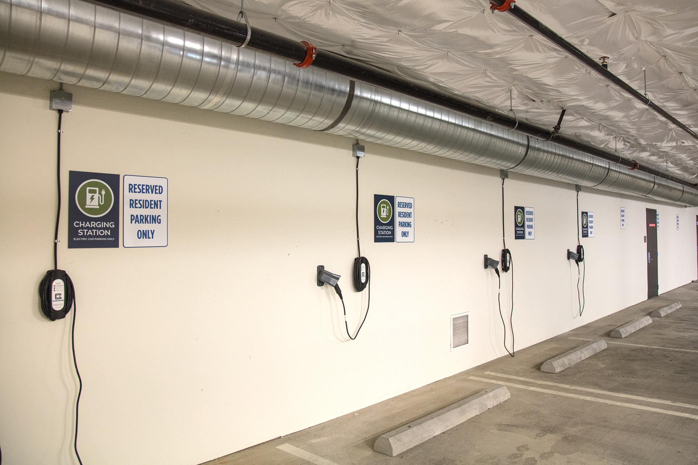 Several electric vehicle charging stations are mounted on a white wall in a parking garage. Signs indicate "RESERVED RESIDENT PARKING ONLY" and "CHARGING STATION." The ceiling has exposed ductwork, and the parking spaces are empty, emphasizing the dedicated area for electric vehicle charging.