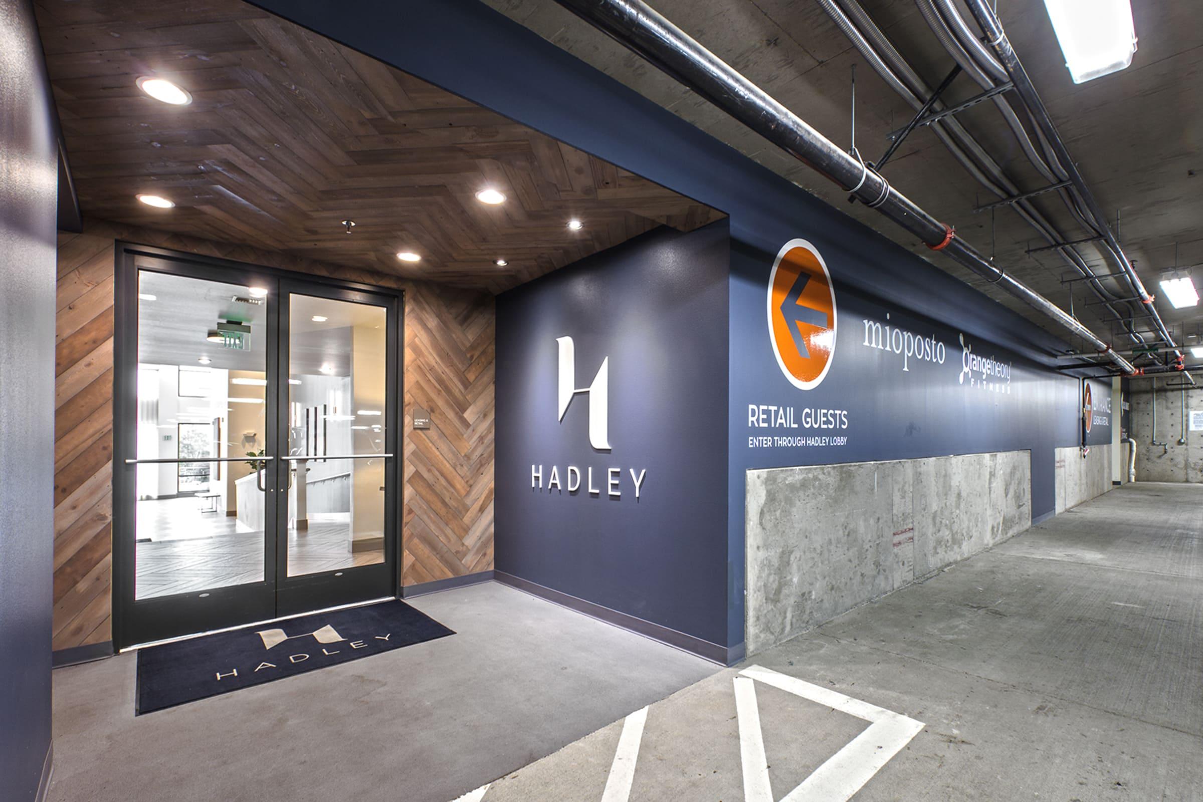 Entrance to Hadley, featuring a stylish design with wooden accents and a modern logo. A sign indicates the area is for retail guests, leading into a well-lit lobby area. The surroundings are part of a parking structure that complements the upscale ambiance.