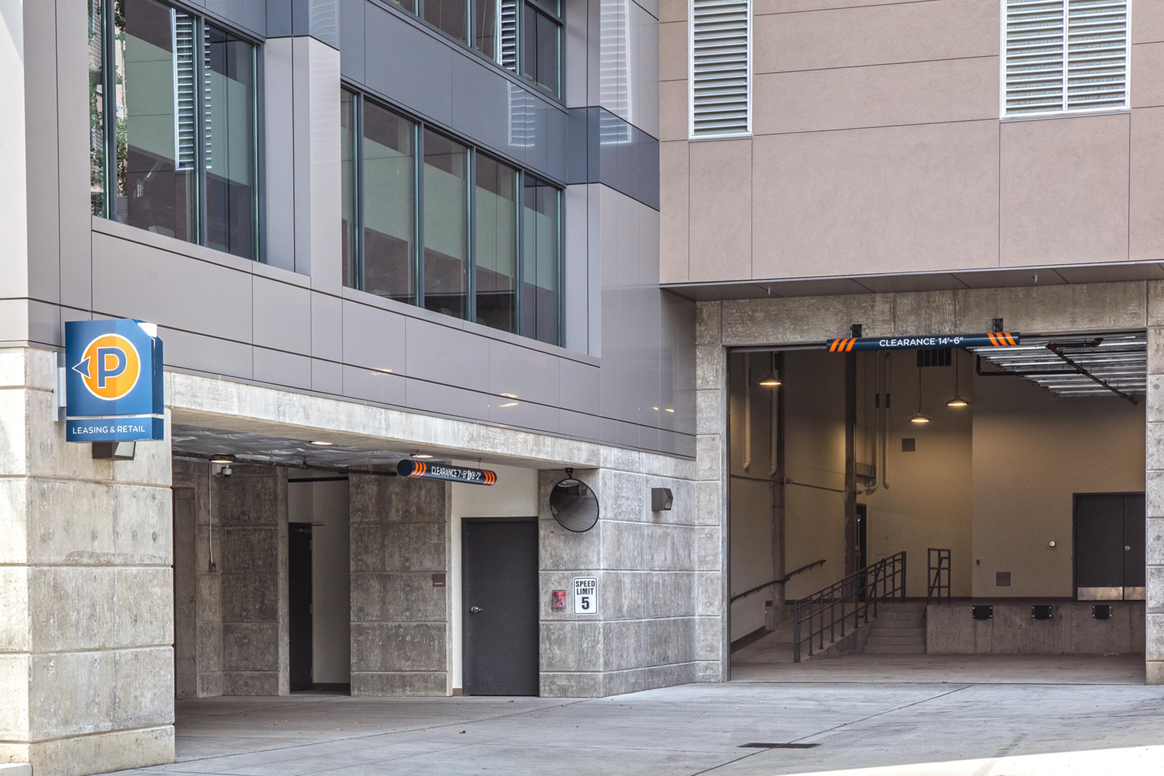 Parking garage entrance with a sign indicating clearance height. On the left, a blue parking sign is visible, and on the right, a ramp leads to the garage entrance, which is surrounded by modern building architecture. The area appears well-maintained with concrete and glass design elements.