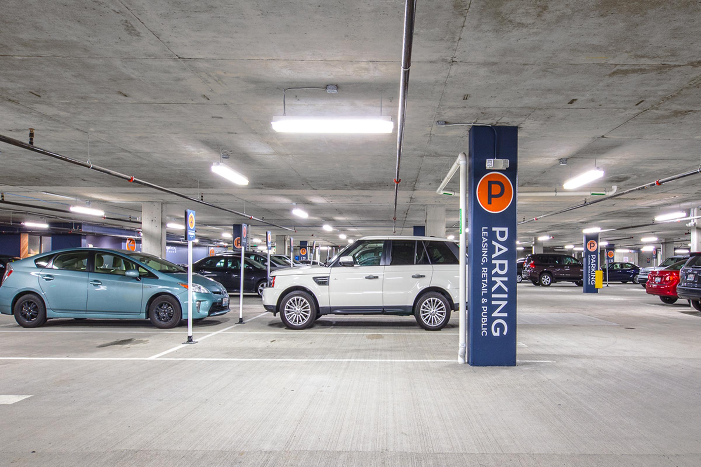 A spacious indoor parking garage with multiple parked vehicles. A white SUV occupies a spot next to a teal car. Bright overhead lights illuminate the area, and clear signage indicates parking rules. The floor is concrete, and several other cars are parked in the background.