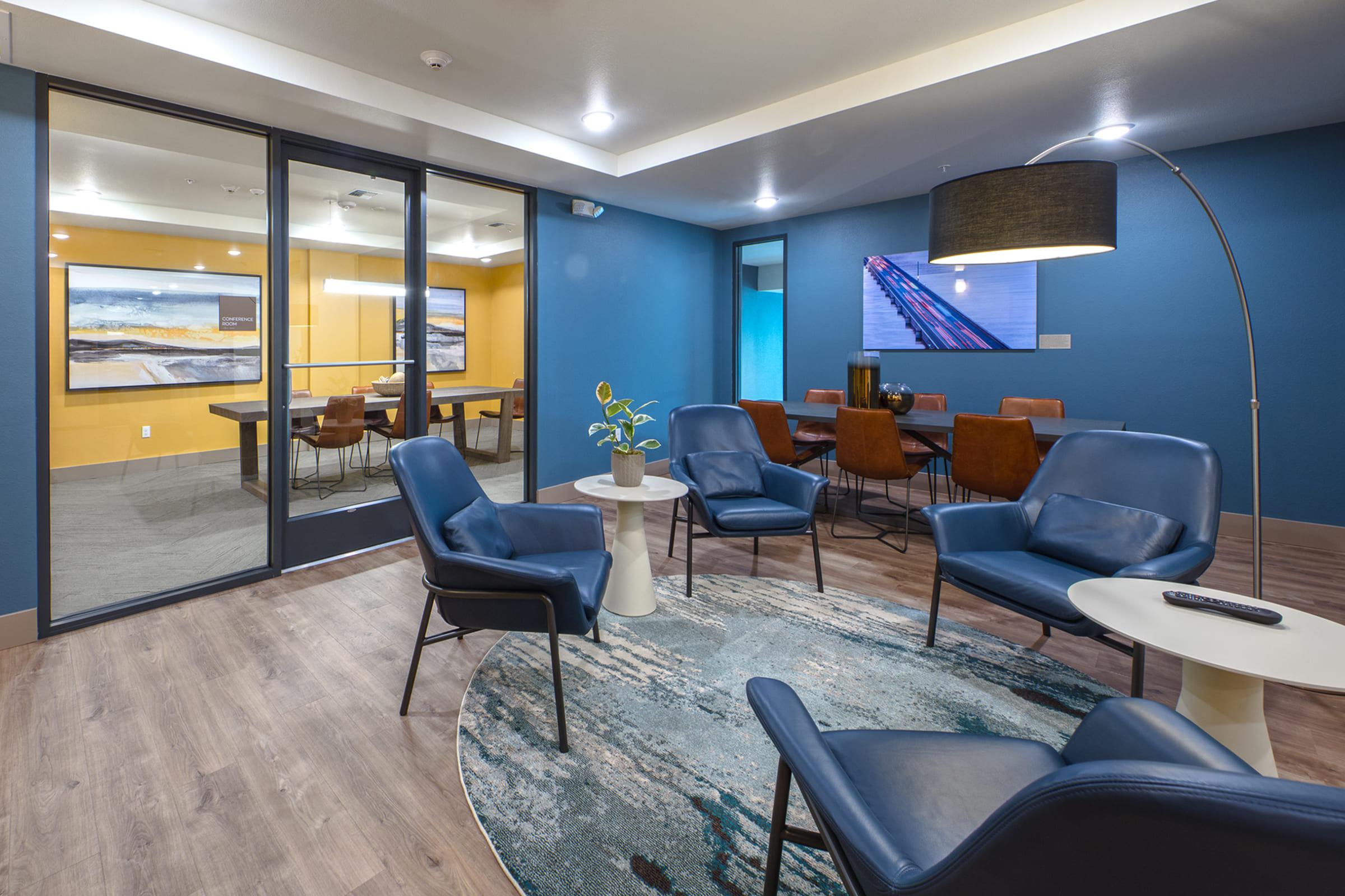 A modern interior space featuring a sitting area with blue chairs arranged around a round table, a decorative plant, and a stylish floor lamp. The walls are painted blue with artwork on display. In the background, a conference room is visible through glass doors, showcasing additional seating and a bright yellow wall.