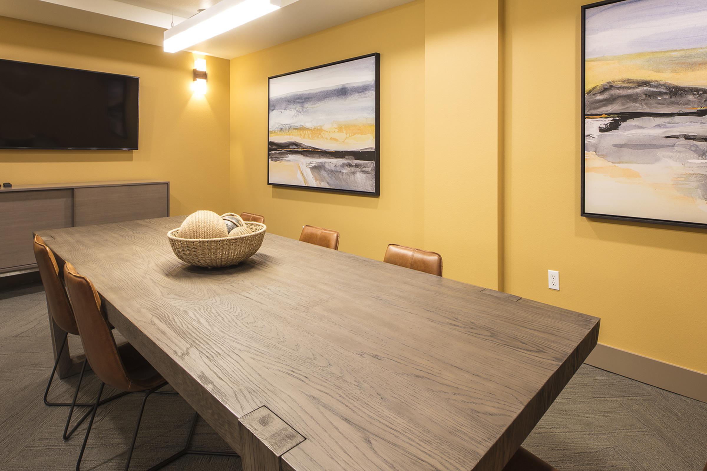 A modern conference room featuring a long wooden table surrounded by leather chairs. A decorative basket sits in the center of the table. The walls are painted a warm yellow, and there are two abstract paintings hanging on the wall. A large screen is mounted on one wall.