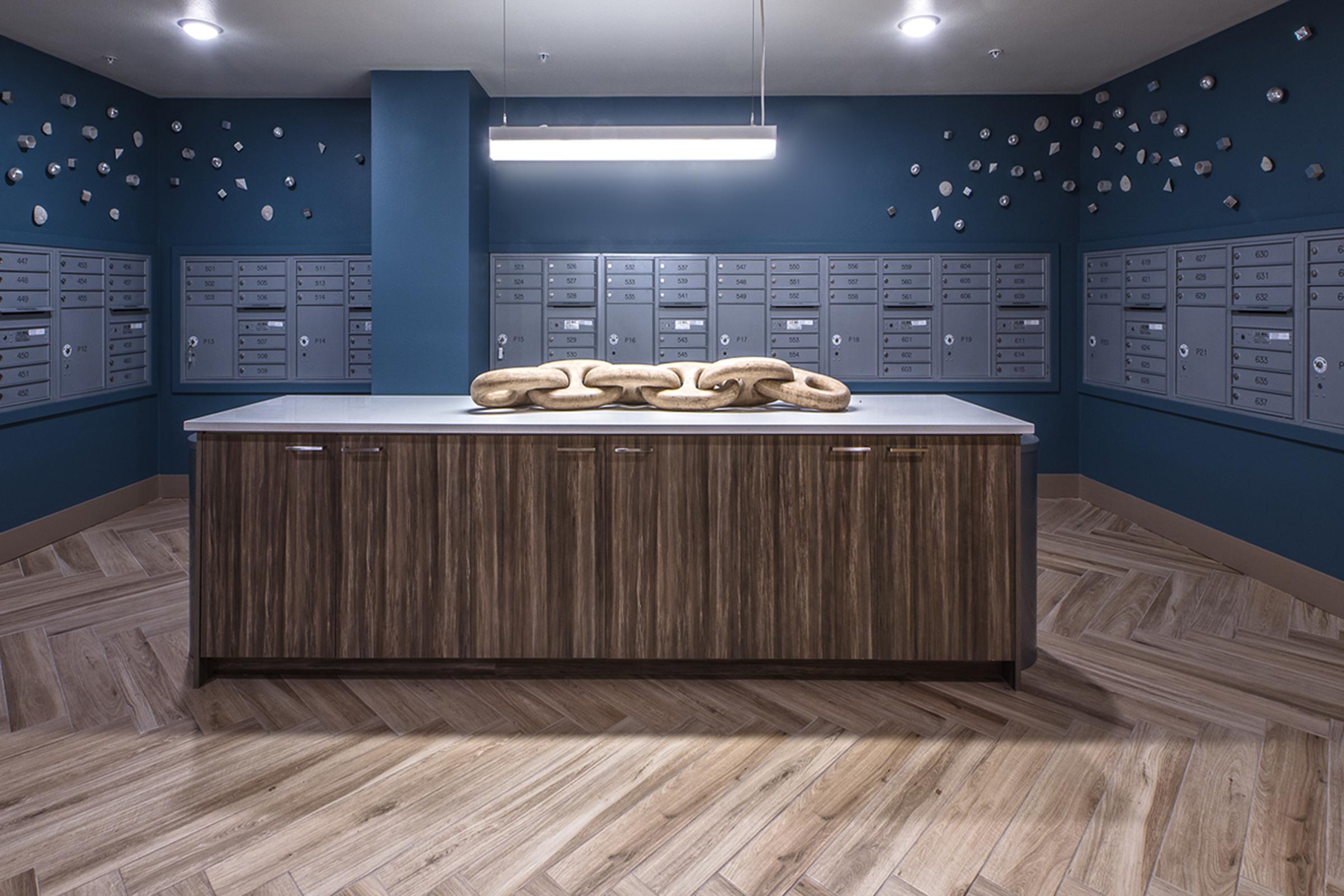 Interior of a modern mailroom featuring a sleek wooden counter and numerous gray mailboxes on the walls. The background is painted dark blue, and decorative elements are scattered around the room. The flooring consists of herringbone-patterned wood, creating a warm, inviting atmosphere.