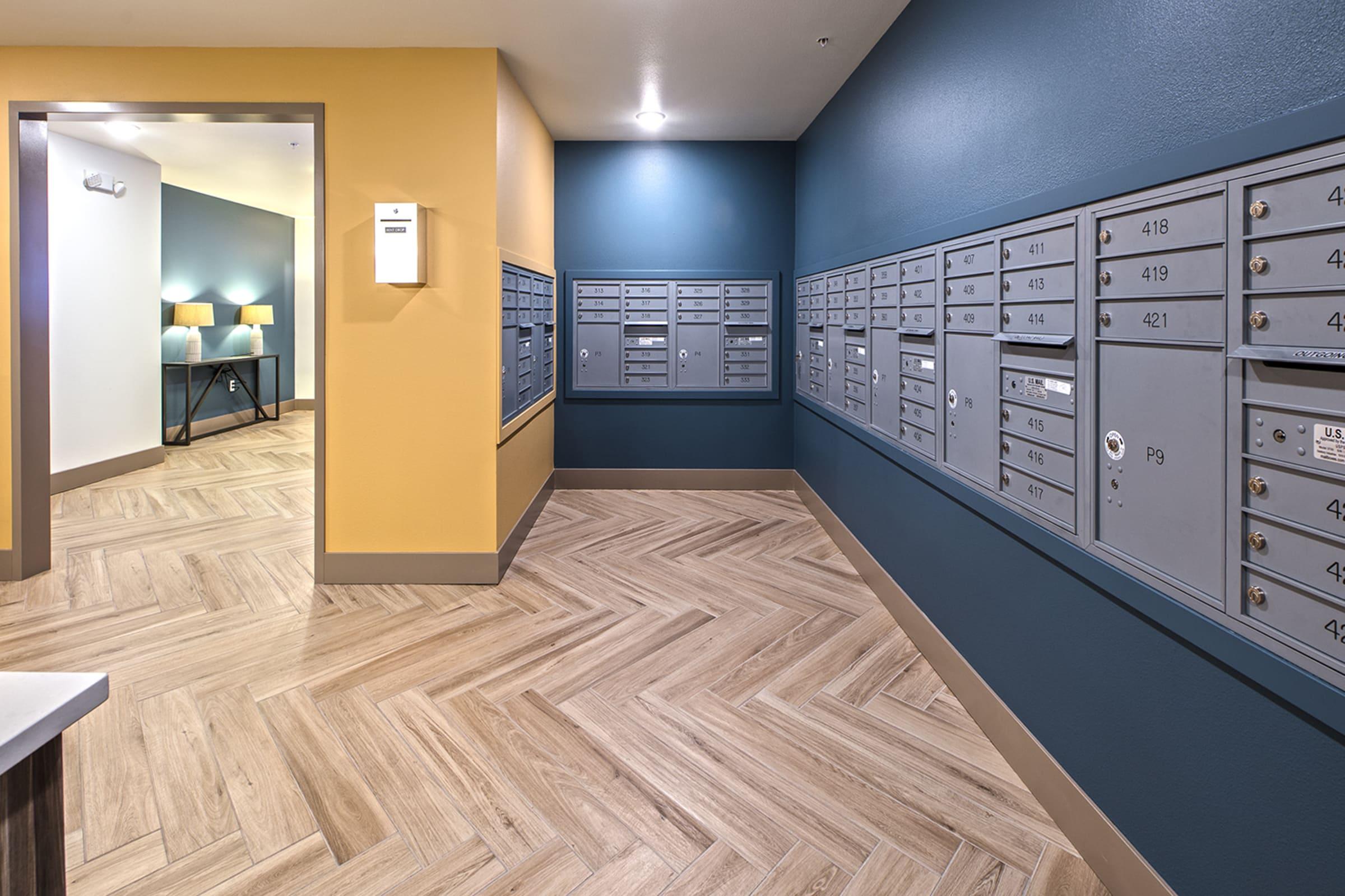 Interior view of a mailroom featuring a series of gray mailboxes affixed to the wall. The space has a warm color scheme with teal and yellow walls, and a wooden floor with a herringbone pattern. A doorway leads to another room brightly lit with two lamps.