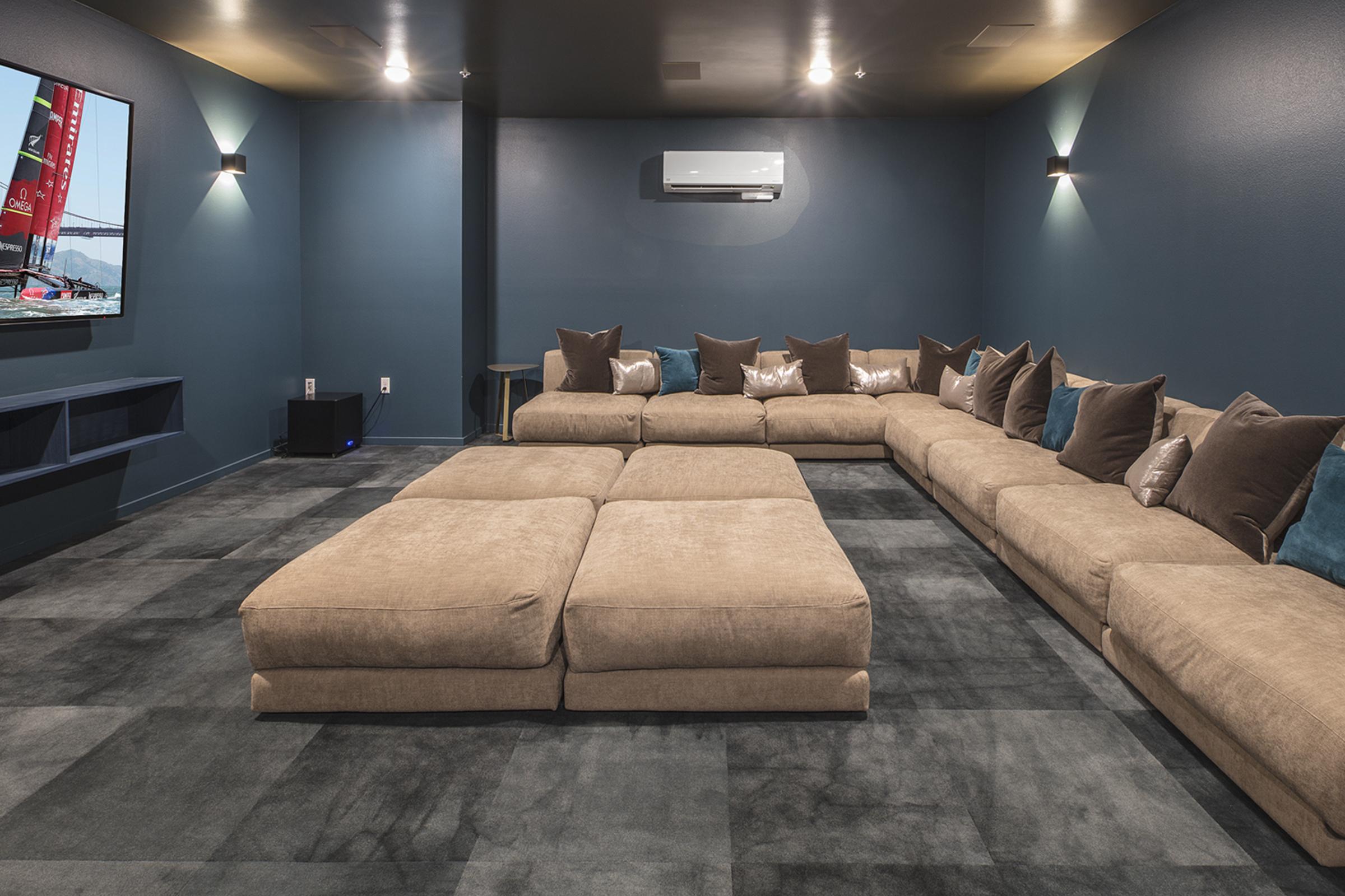 A cozy home theater room featuring a large, L-shaped sectional sofa with numerous pillows, a flat-screen TV mounted on the wall, and ambient lighting. The walls are painted in a deep blue color, and the floor has a modern gray carpet, creating a comfortable and inviting atmosphere for watching movies.