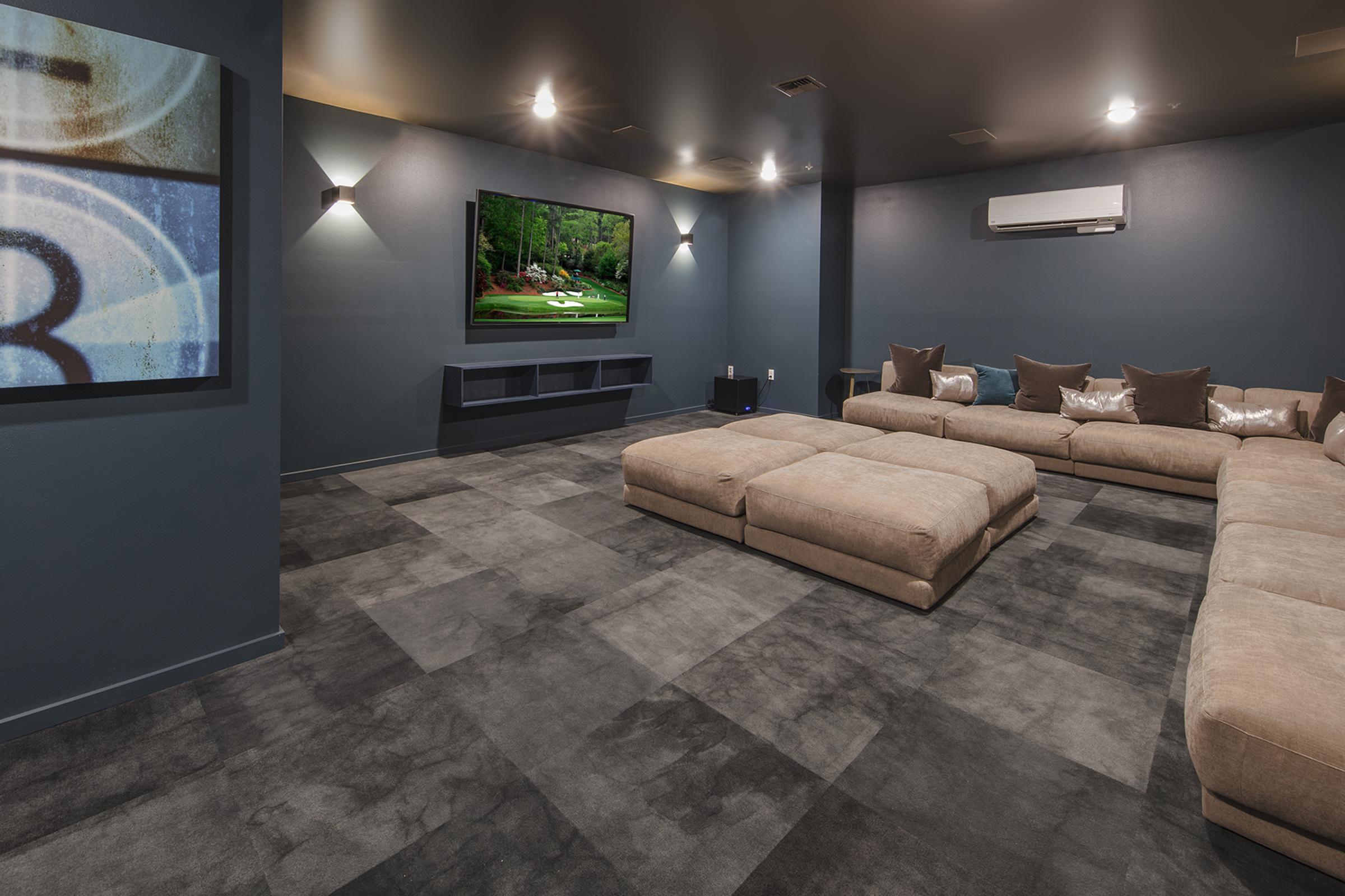 A cozy home theater room featuring a large flat-screen TV mounted on the wall, soft beige sectional seating, and decorative pillows. The walls are painted a deep blue hue, and the floor is covered with gray carpet tiles. Subtle lighting highlights the space, creating a comfortable ambiance.