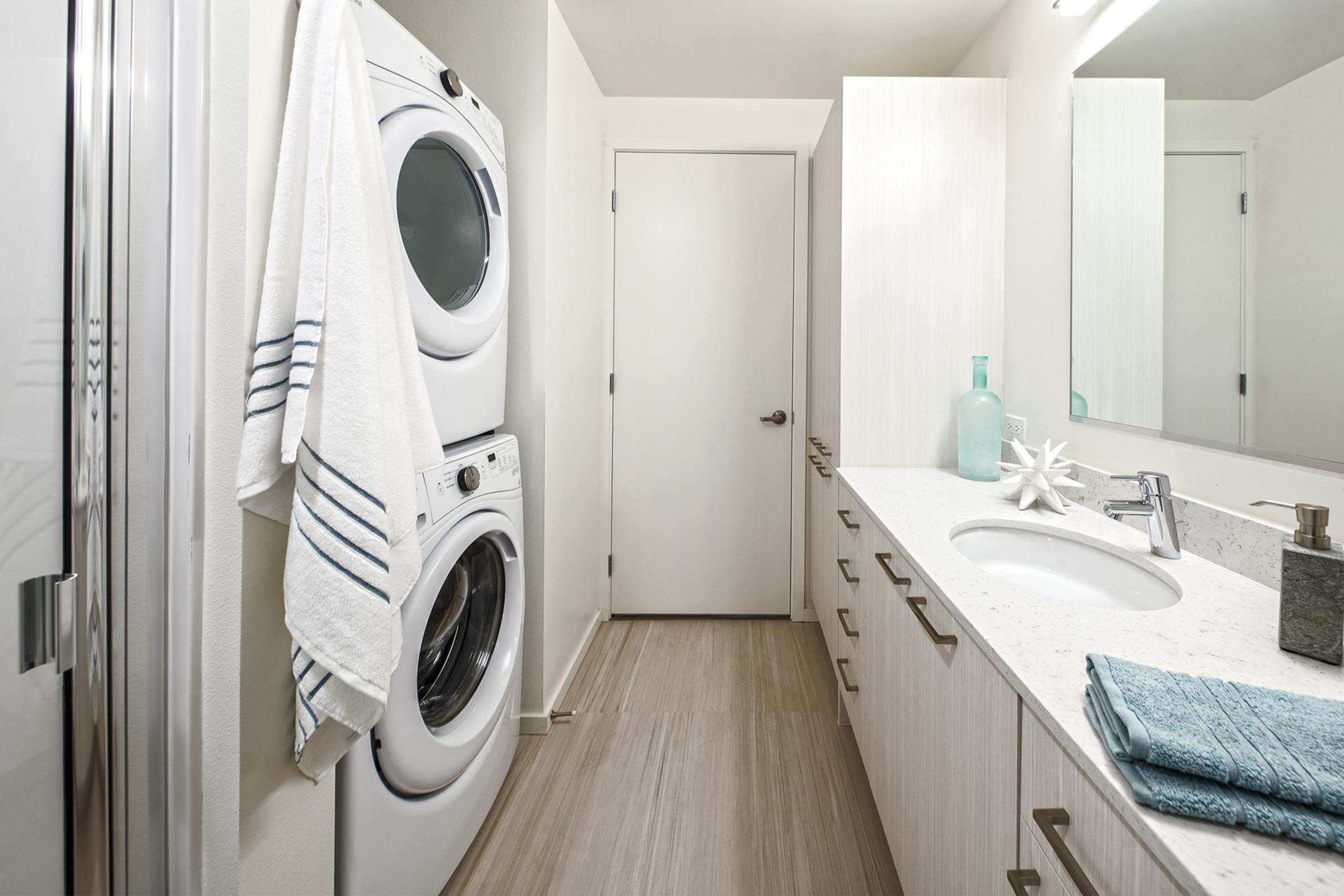 A modern laundry room featuring a stacked washer and dryer, a white countertop with a sink, and cabinets.